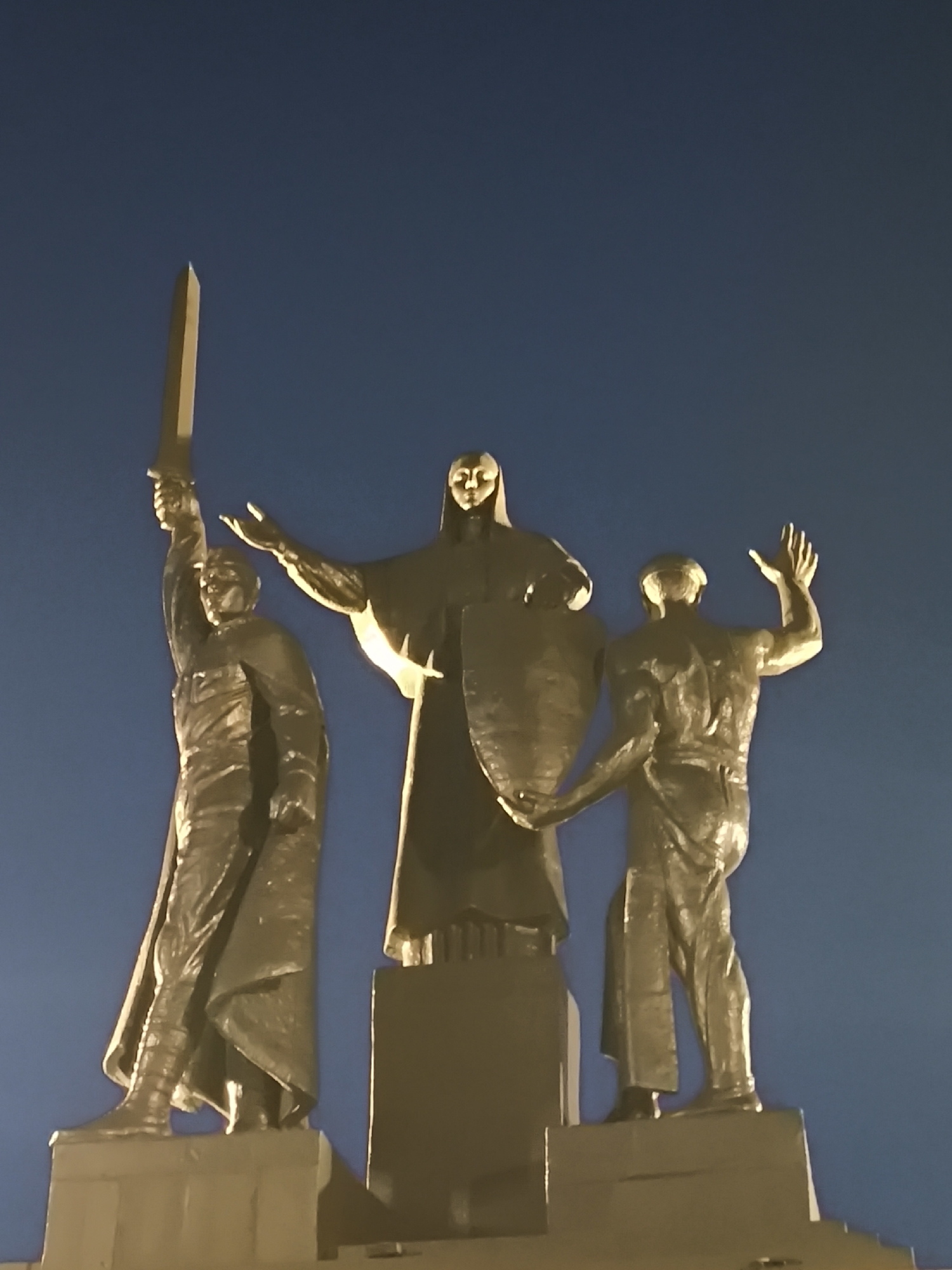 The image shows a bronze sculpture at night against a dark blue sky. The sculpture depicts three figures on pedestals. The central figure is a woman in a robe, holding a shield and extending her left arm. To her left stands a male figure, also in bronze, raising a sword with his right hand. To the woman's right is another male figure in bronze, with his right arm raised in a waving gesture. The sculpture is illuminated, casting a warm glow on the figures. This monument is the "Perm Gate" or "The Great Perm," located in the city of Perm, Russia.