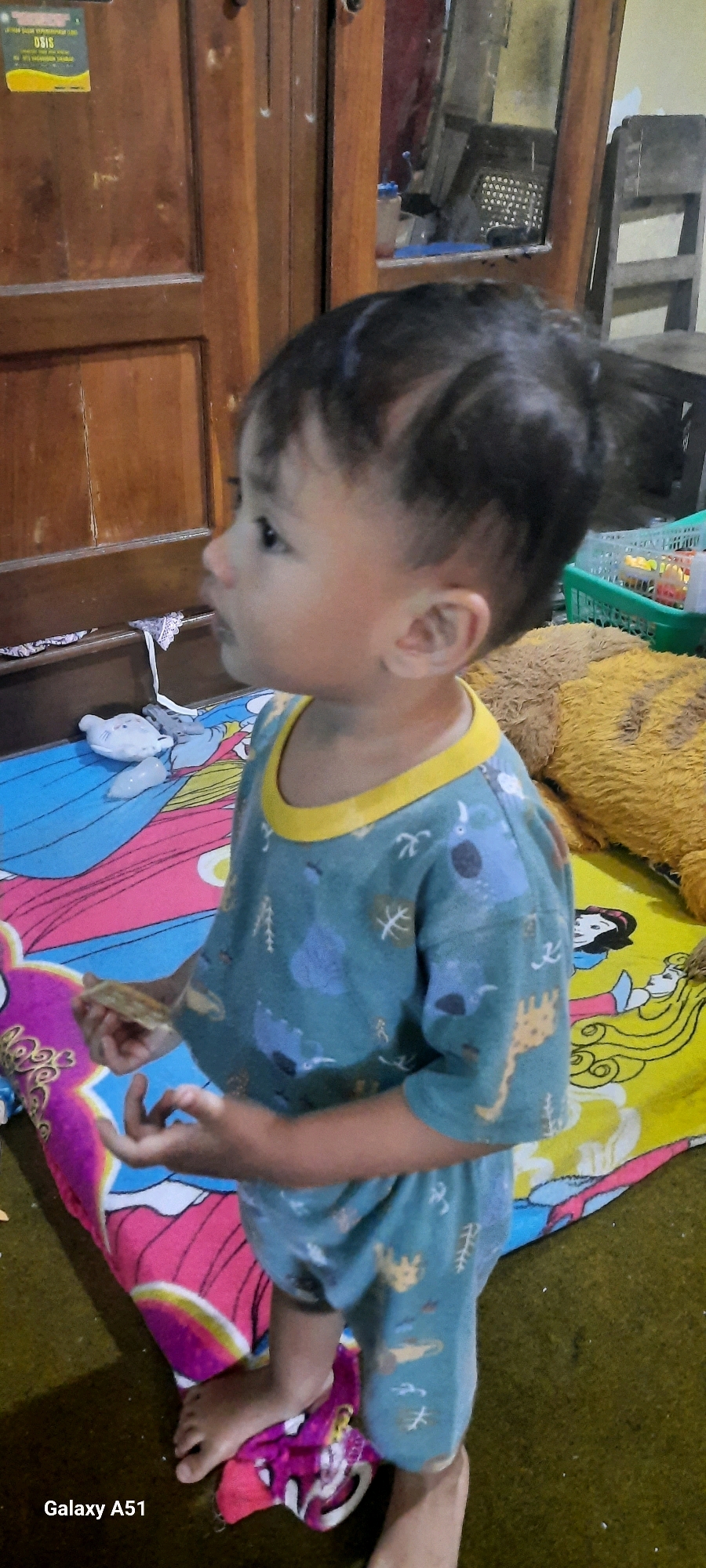 In the city of Jatitengah, Indonesia, a young child is standing on a colorful blanket, wearing a blue t-shirt and shorts with a pattern of various animals and trees. The child is holding a small, flat object in their right hand and looking to the left. The blanket is spread on the floor, and behind the child is a wooden cabinet. In the background, a large stuffed animal and a green plastic basket are visible. The lighting suggests an indoor setting, possibly a home.