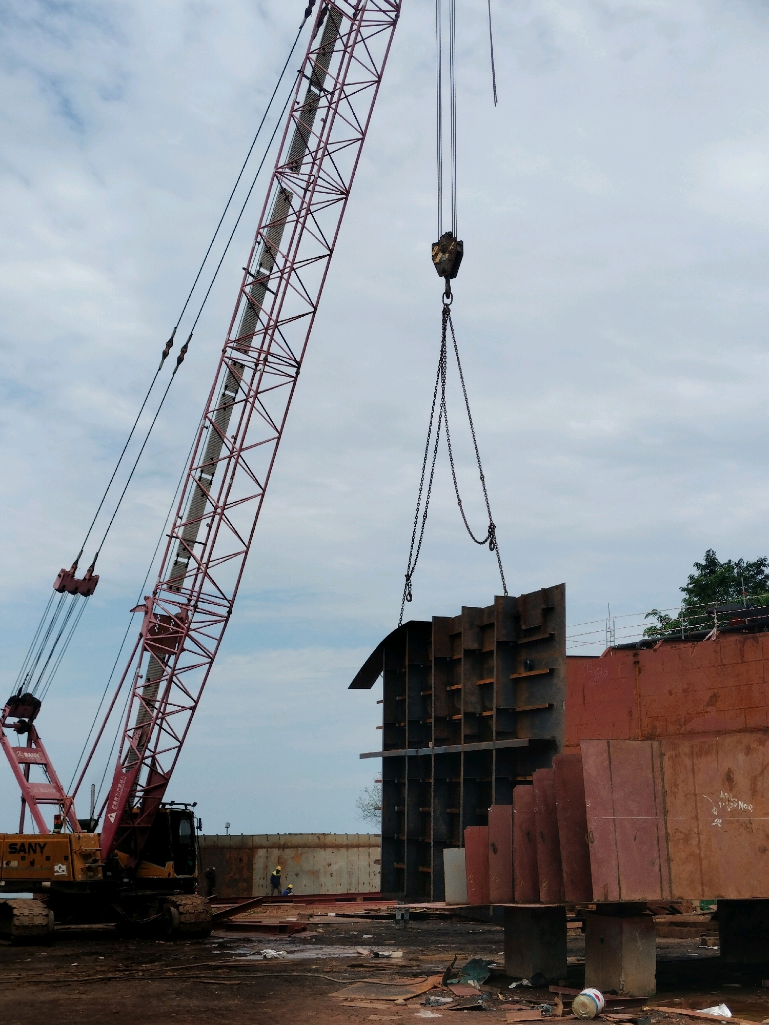 A large, red and yellow SANY brand crawler crane is actively engaged in lifting a section of a large, prefabricated metal structure. The crane's boom extends diagonally from the lower left towards the upper center of the frame. The object being lifted is a dark gray, skeletal metal piece with multiple vertical and horizontal supports, suspended by thick chains and a hook. To the right of the lifted structure, a large, rust-colored metal wall, likely part of a ship or industrial building, is partially visible. Several workers in yellow hard hats are present in the background, near a long, segmented metal wall that appears to be under construction. The ground is covered in dirt, debris, and various metal scraps, suggesting an active industrial or shipyard environment. The sky above is overcast with scattered clouds. This scene is likely located in a shipyard or heavy industrial area within Batam, Indonesia, known for its shipbuilding and manufacturing industries.