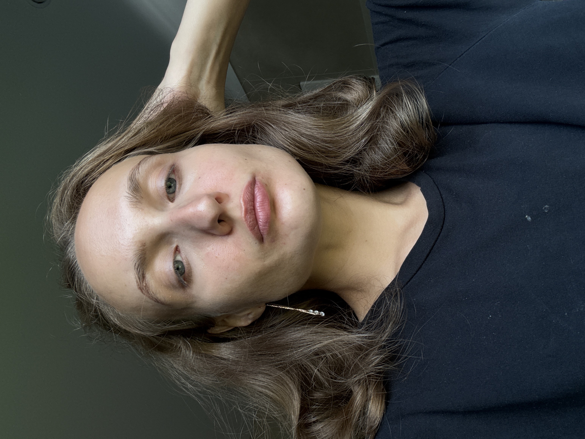 A young woman with shoulder-length wavy brown hair is shown from the chest up. She is wearing a black t-shirt. She is looking forward with her right hand touching her head. She has light skin, light blue eyes, and light eyebrows. She is wearing a long gold earring with three small clear crystals. The background is a dark, out-of-focus wall. There is no indication of the location being in the city of Подшивалово, Russia.