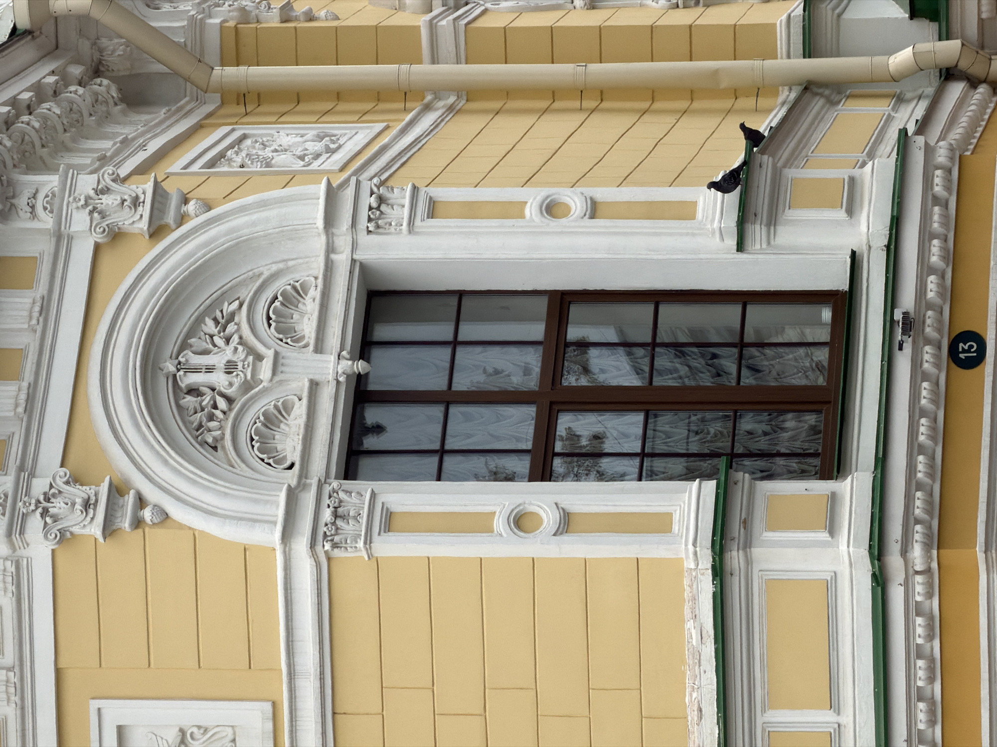 This is a close-up, high-angle, outdoor, eye-level shot of a building facade with ornate white stucco decorations against a yellow wall. The building appears to be in Nizhny Novgorod, Russia. Sunlight illuminates the scene, suggesting it is daytime.
The main subject is the architecture. A large, arched window with dark brown framing and multiple panes dominates the center of the image. The window is framed by intricate white decorative elements, including volutes, acanthus leaves, and geometric patterns. Above the window, a cream-colored pipe runs horizontally across the building, a section of which is bent at an angle.
Two pigeons are perched on the white architectural ledge above the window, one facing left and the other facing right. They appear to be resting. The yellow wall is textured with vertical lines, creating a subtle ribbed effect.
To the right of the window, a green vertical element is visible, possibly part of the building’s trim or a downspout. Further to the right, a circular emblem with the number "13" in white is affixed to the yellow wall, likely a building or apartment number.
The overall impression is one of classical or baroque architecture, characterized by its elaborate ornamentation and the contrast between the bright yellow and the detailed white stucco. The scene is static, with no discernable activity beyond the pigeons' presence.