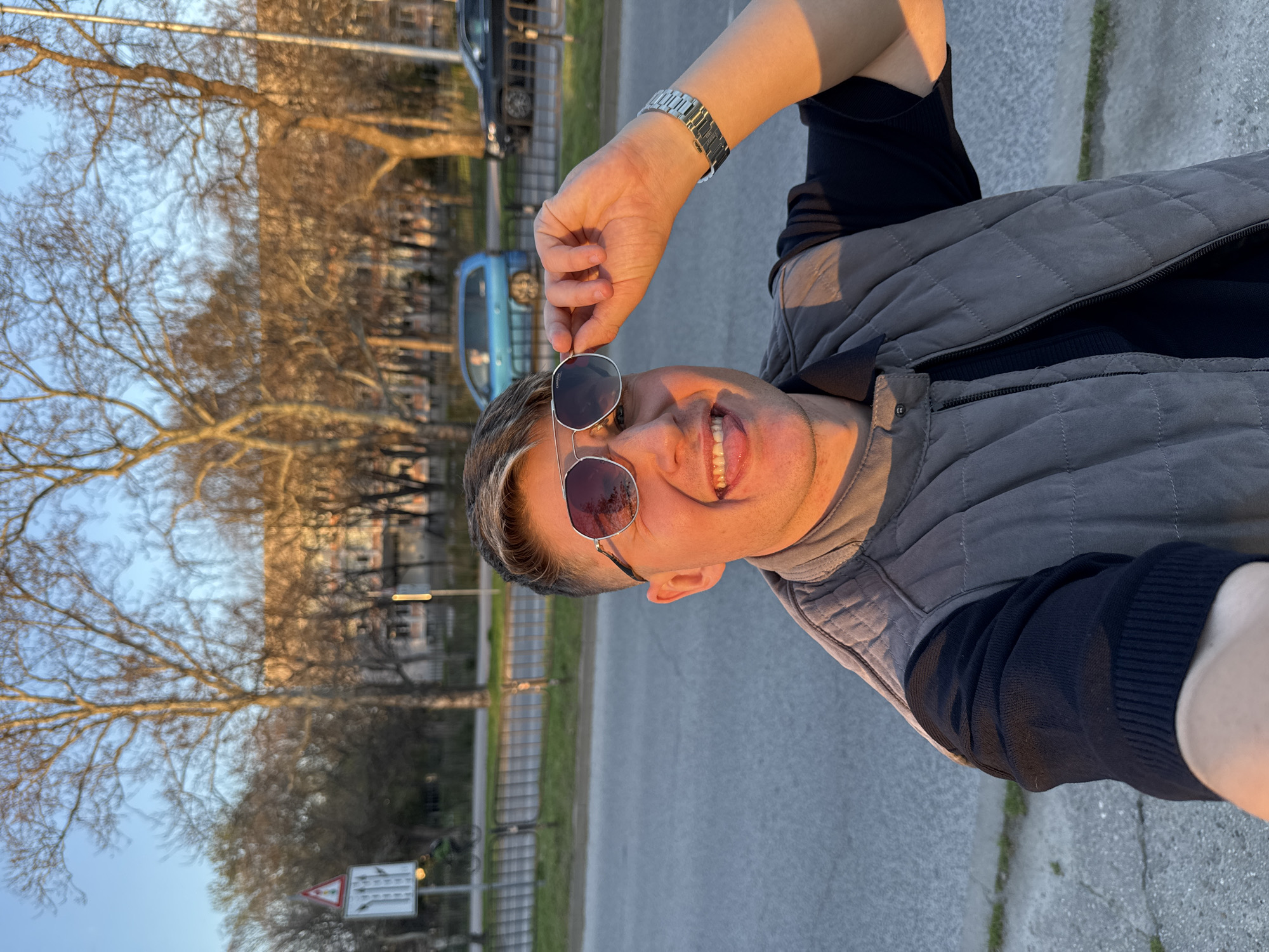 A fair-skinned individual with short, styled hair poses for a selfie outdoors. The person is wearing sunglasses, a black long-sleeved shirt, and a quilted gray vest. They are holding the sunglasses in one hand, partially obstructing their face, and smiling with their tongue partially visible. A metal wristwatch is visible on their wrist.
The background includes a street with a sidewalk, grass, and trees without leaves. Cars are parked along the street. Based on the architectural style and general appearance, the location is consistent with urban areas in Plovdiv, Bulgaria, though without specific landmarks, a precise location is not determinable. The time appears to be late afternoon based on the warm, golden lighting.