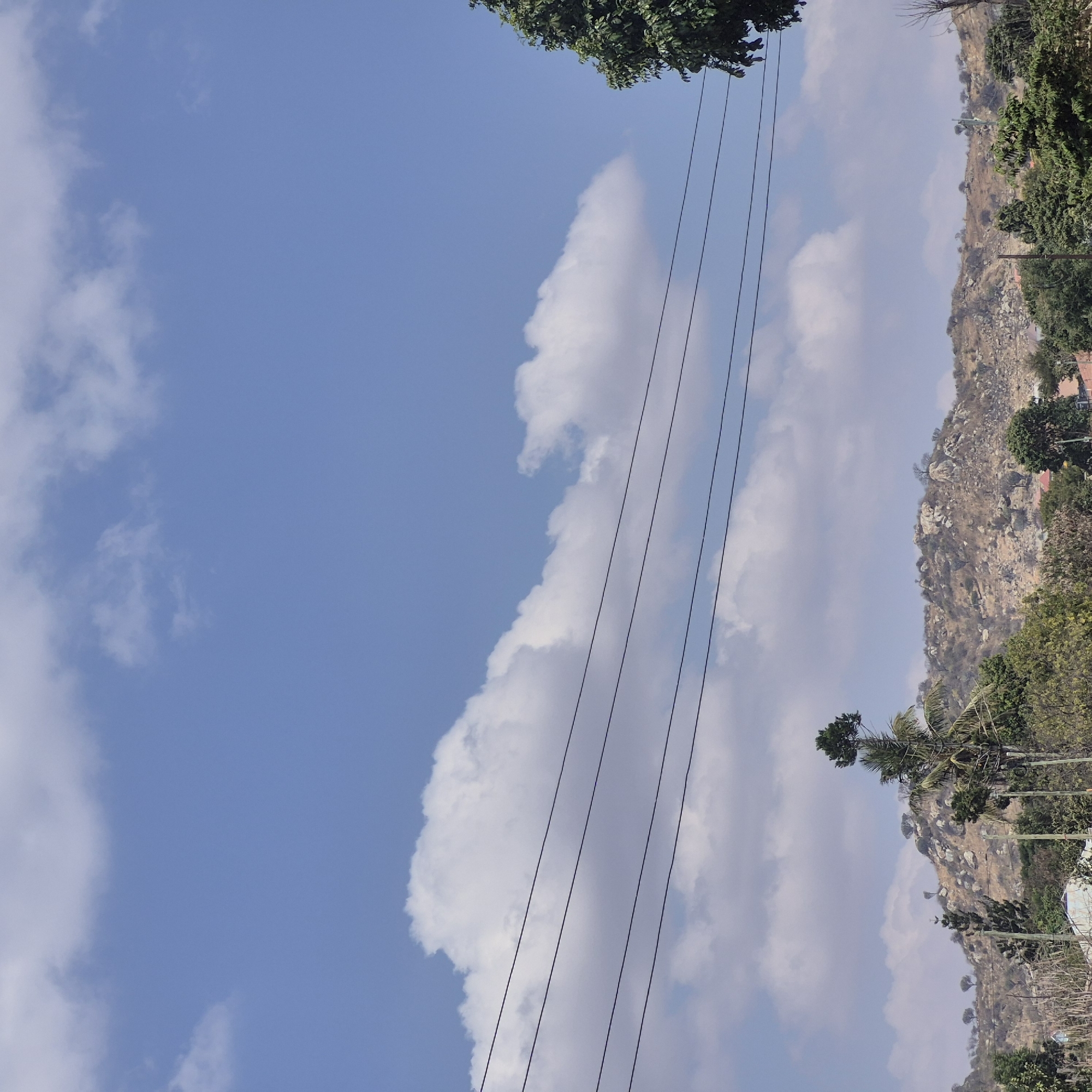 The image depicts a bright, daytime scene with a clear blue sky dotted with fluffy white clouds. In the foreground, four dark electrical wires stretch horizontally across the frame. Beneath the wires lies a rugged, rocky hillside covered in sparse, dry vegetation and scattered trees. A few buildings are visible amongst the trees at the base of the hill. The landscape suggests a semi-arid or tropical environment. The specific location is identified as Dodoma, Tanzania, an area known for its hilly terrain and dry climate. There are no people or animals visible in the image.