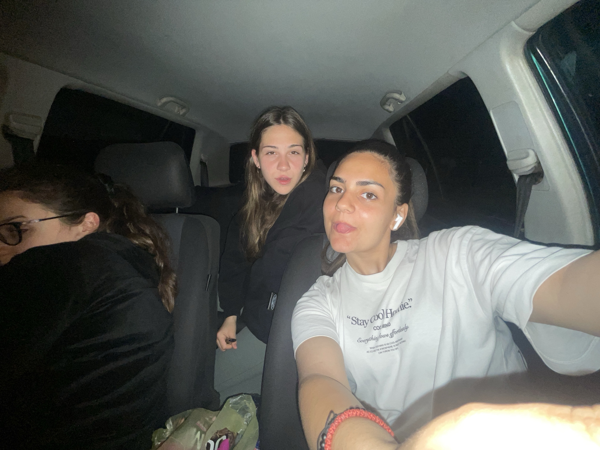 Three women are inside a car at night. The car is stationary. The woman on the right is taking a selfie, sticking her tongue out. She is wearing a white T-shirt with the text "Stay cool Hoodie." She has an earpod in her ear. The woman in the middle is looking at the camera. She is wearing a black hoodie. The woman on the left is wearing glasses and has her head turned away from the camera. The environment is dark. The city of Debelt, Bulgaria, is not discernible.