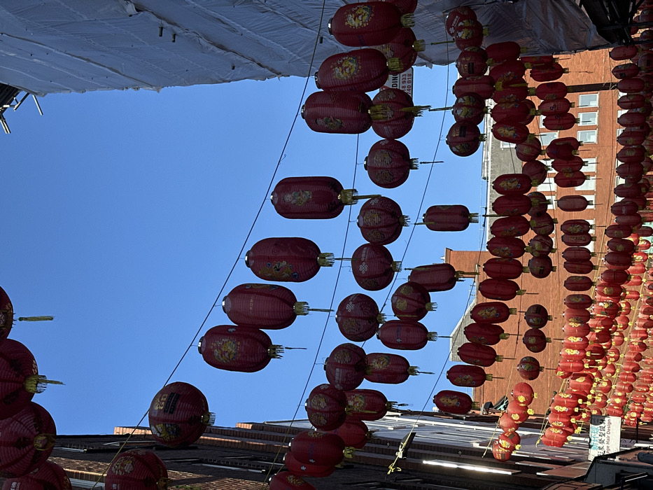 The image depicts a narrow street in Chinatown, London, United Kingdom, viewed looking upwards towards a clear, bright blue sky. Hundreds of red Chinese lanterns are densely suspended on black wires strung horizontally across the street at various heights. The lanterns are primarily cylindrical or oval-shaped, with some spherical, and feature gold decorative elements including Chinese characters and intricate patterns.
On the left side, a multi-story brick building with windows is partially visible, its facade partially shaded. Towards the bottom left, a faint sign with Chinese characters and the words "Hair Design" is discernible. The right side of the street features a tall building completely enveloped in white protective sheeting, indicative of ongoing construction or renovation, with scaffolding elements visible at the top. A white sign attached to this covered building displays partial text, including "DING" and phone numbers such as "078 933" and "0 740 018". Further down the street, red brick buildings with a pitched roof are visible in the background. No individuals are prominently visible within the frame.