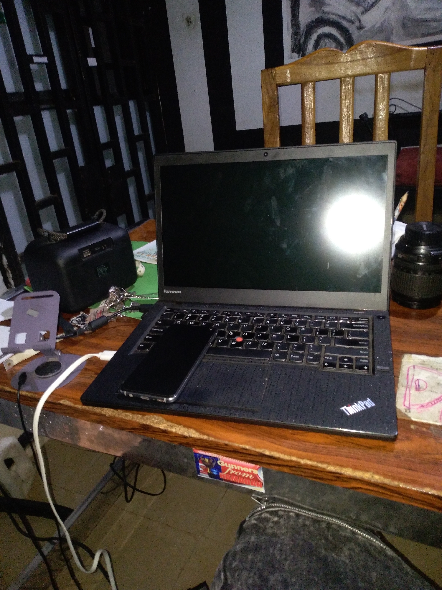 A Lenovo laptop is open on a wooden desk. A black smartphone is placed on the laptop's keyboard. To the left of the laptop, there is a black portable speaker and a grey phone stand with a white charging cable. A black camera lens is to the right of the laptop. The background features a striped wall with artwork and a wooden chair. The scene is lit by artificial light, creating reflections on the laptop screen.