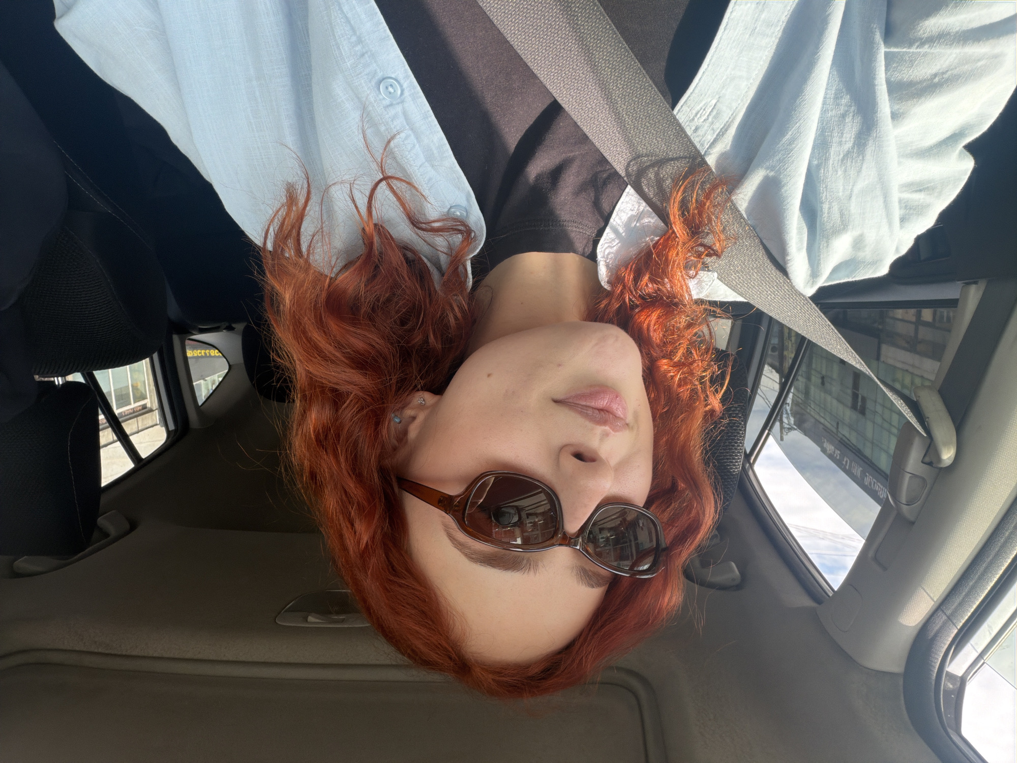 This media file is an upside-down selfie of a person with long, wavy, bright red hair, seated inside a car on a sunny day. The person is wearing dark brown oval sunglasses, a light blue button-down shirt layered over a dark top, and a grey seatbelt across their chest. A small blue stud earring is visible in their left ear.
The car's interior is visible, including the light-colored headliner, a black headrest, and parts of the door panel and window frames. The bright daylight creates strong shadows and reflections. Reflections in the sunglasses show an indoor environment with shelves, possibly from a store or building the car is parked near.
Through the car windows, external views are visible. On the left side, a blurry building and what appears to be a street or parking lot can be seen. On the right, a large, modern building with numerous windows reflects the bright sky. Blurry text is visible on the side of this building. Closer examination reveals fragments of Georgian text, including `სსაფლავი` (which translates to "cemetery" or "grave," often found in place names or historical markers) and `შტრ. რადგან` (parts of words, "str." could be an abbreviation, or part of a longer word). This visible text, combined with the provided location context, places the scene in Tbilisi, Georgia. The image conveys a casual moment captured during daylight hours from within a vehicle.