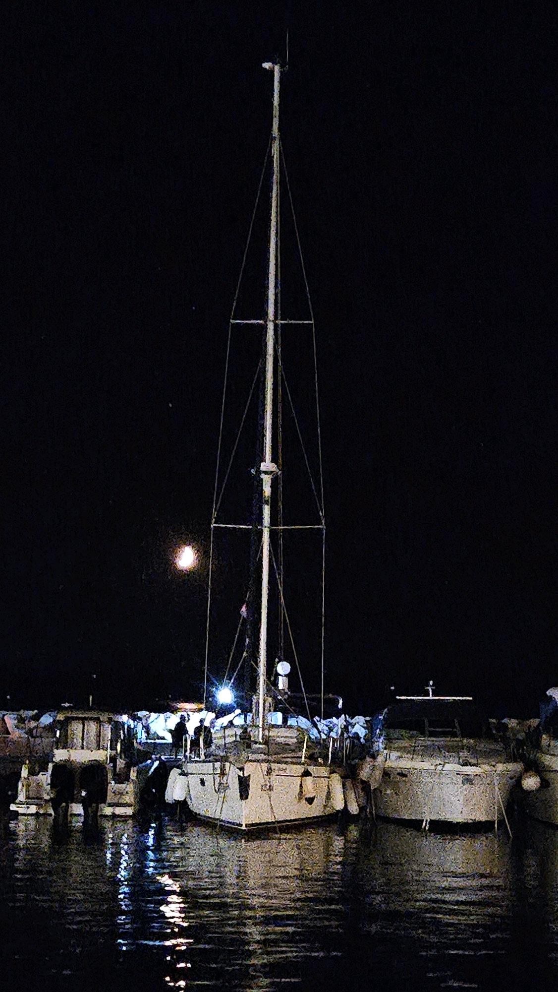 A tall sailboat mast dominates the center of the image against a dark night sky, illuminated by a crescent moon. The sailboat is docked at night, reflecting in the rippling water. Several other boats are docked alongside it. In the background, there are figures on the deck of the sailboat and adjacent structures, some of which are lit, casting reflections on the water. The scene appears to be a marina in Općina Tučepi, Croatia.