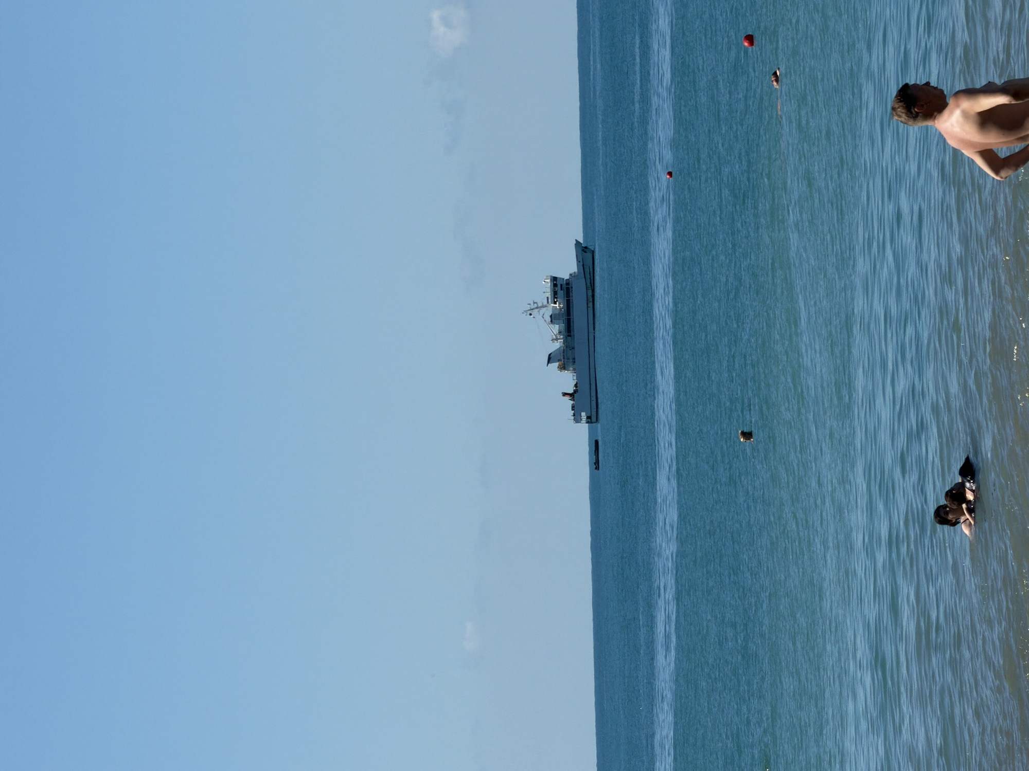 The image depicts a serene coastal scene under a clear blue sky, presumably on a warm day. In the background, a white ship with several decks and a superstructure is visible sailing on the calm, turquoise sea. The ship appears to be a ferry or a similar vessel. In the foreground, several people are in the water. Close to the viewer, a man with his back to the camera is standing in shallow water, looking out towards the sea. Further out, two people are swimming together, one appearing to be an adult with a child. Another individual's head is visible as they swim in the distance. Red buoys are scattered across the water, suggesting a designated swimming area or navigational markers. The water is slightly rippled, indicating a gentle breeze. The overall atmosphere is one of relaxation and recreation. The presence of a large ship on the horizon suggests this location is a coastal city with maritime activity, and given the prompt's mention, likely Sochi, Russia.