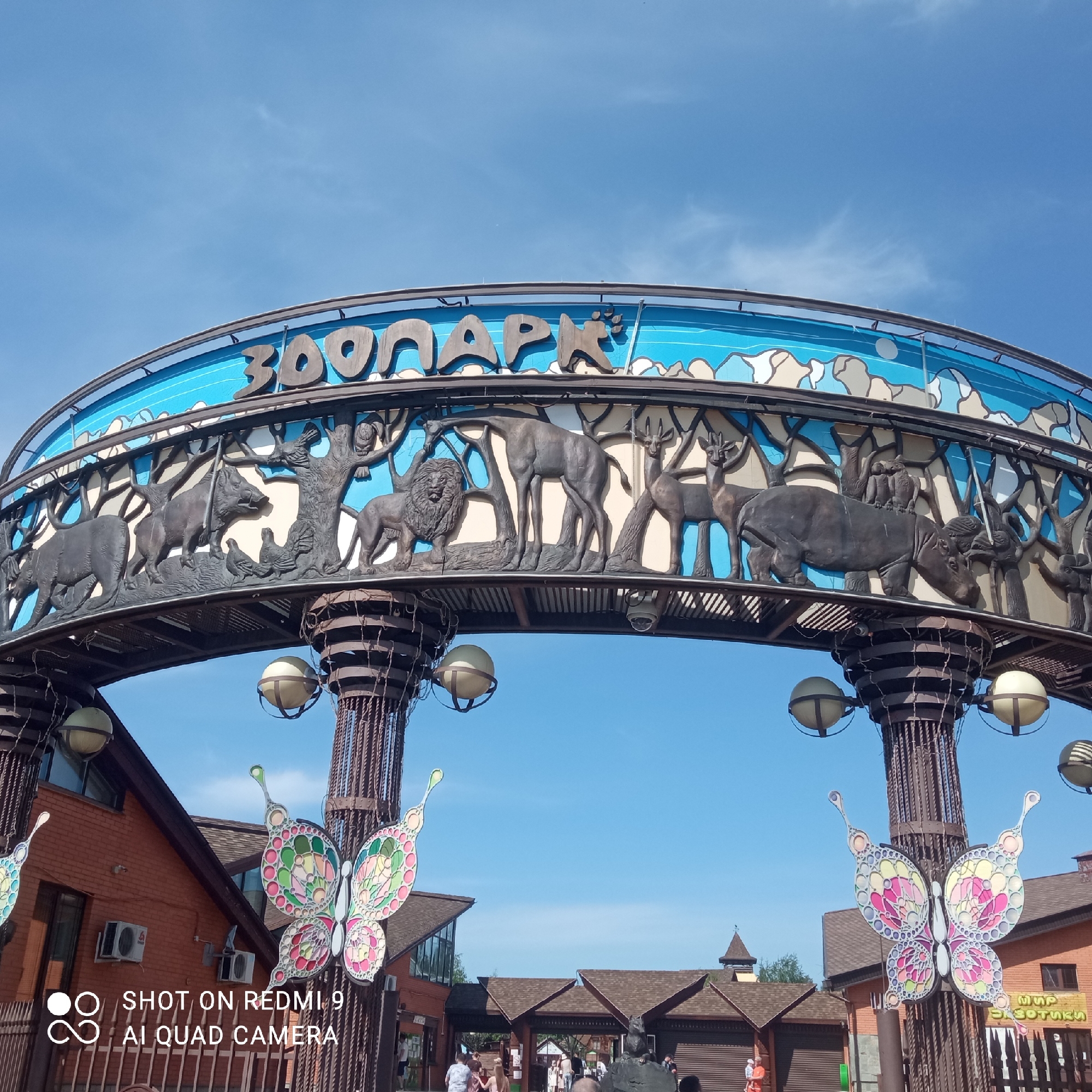 The image captures the entrance arch of the Izhevsk Zoo in Russia. The arch is decorated with animal sculptures including a rhinoceros, lion, deer and hippo, arranged among tree figures along the archway. The Russian word for zoo, "ЗООПАРК" is written across the top of the arch in large, stylized letters, set against a light blue background that depicts stylized water and light-colored clouds. Supporting the arch are two pillars, each adorned with spherical lights. Large butterfly decorations are attached to the pillars. The background includes brick buildings and a clear, blue sky. Several people are visible passing through the entrance and gathering near the entry area.