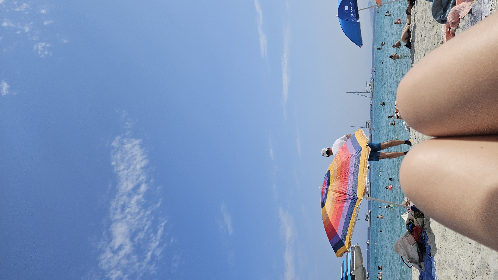 This is a first-person perspective photo taken on a sandy beach in L'Île-Rousse, France. The foreground is dominated by the thighs of the person taking the photo, with the beach sand and a striped towel visible just below. In the middle ground, a person wearing a white t-shirt and blue shorts stands near a colorful, striped beach umbrella, facing the water. The sea is calm and clear, with numerous people swimming and a few sailboats in the distance. Another blue umbrella is visible to the right. The sky is a vibrant blue with a few wispy white clouds. The overall scene depicts a bright, sunny day at the beach.