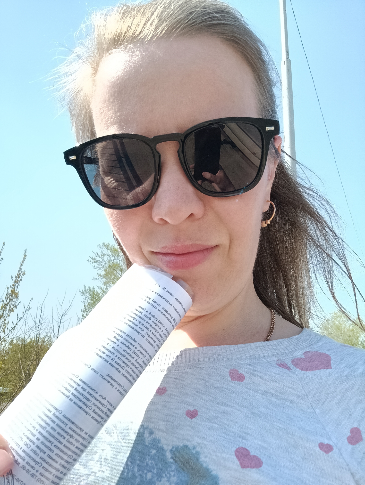 Here is a detailed description of the image:
**People:** The image features a woman, likely in her late 20s or early 30s, with fair skin and light brown hair pulled back. She is wearing black sunglasses, gold hoop earrings, and a light grey sweatshirt with pink heart patterns. A gold necklace is also visible.
**Objects:** A white document with Russian text is held up to her chin. The woman is wearing black sunglasses. There is a utility pole in the background.
**Environment:** The photo appears to be taken outdoors on a sunny day, with a bright blue sky and scattered trees. Based on the text on the document, the location is possibly within the city of Chaikovsky, Russia.