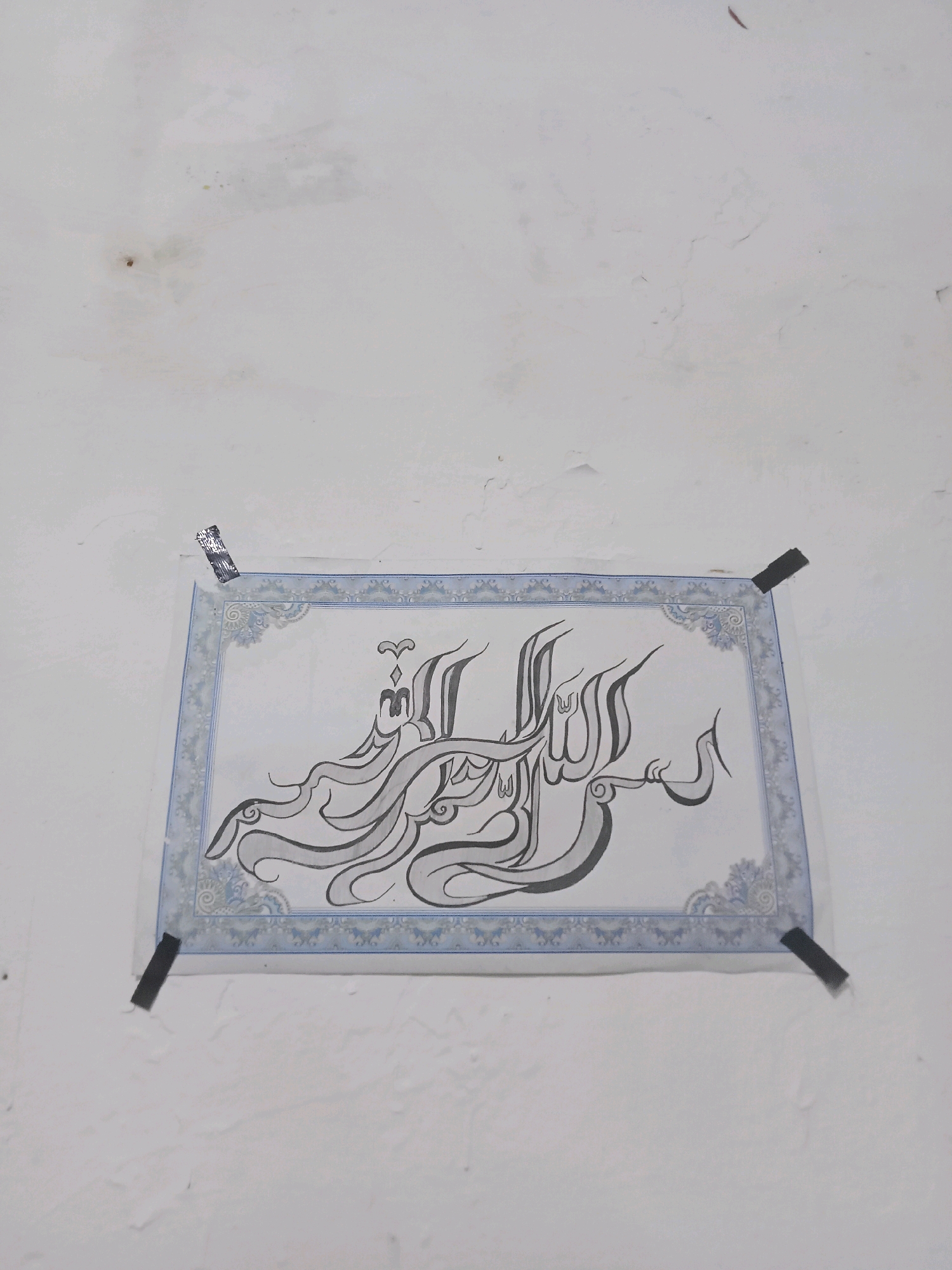 A framed Islamic calligraphy is affixed to a white, textured wall with black tape in the top corners and a silver-patterned piece of tape in the top left. The calligraphy is in black ink with gray shading and appears to be the phrase "Bismillahir Rahmanir Rahim." The frame has a blue and white decorative border. The wall has some discoloration and minor imperfections. The location is identified as Sukomananti, Indonesia.