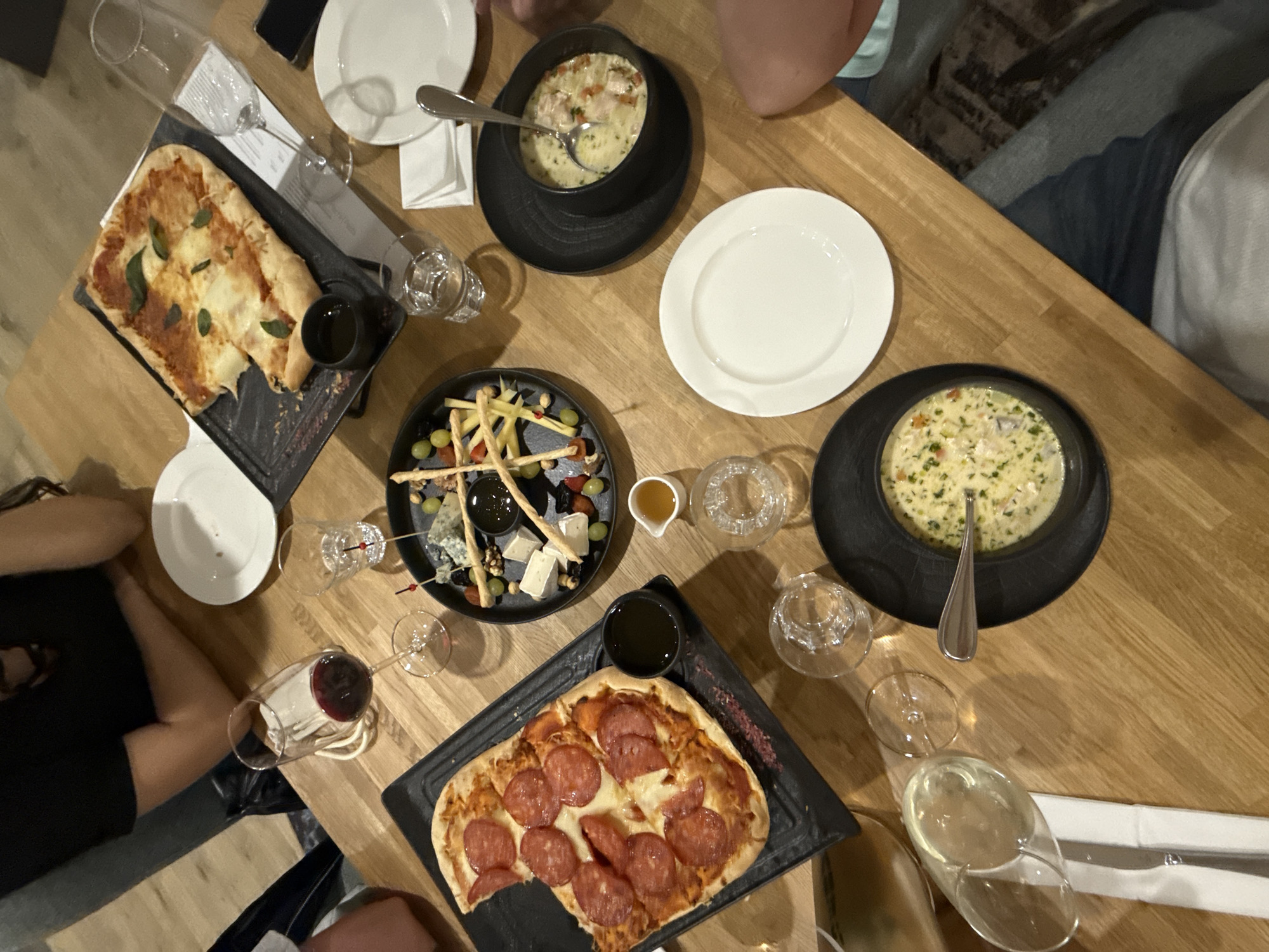 The image shows a wooden table setting for a meal with multiple people. On the table are two bowls of creamy soup in black bowls with spoons, two flatbread pizzas on black platters (one with pepperoni, one with other toppings), a charcuterie board with cheese, grapes, nuts, and breadsticks, a small dish of honey, several wine glasses (one with red wine, one with white wine), and several small glasses filled with water. There are white plates, napkins, and what appears to be a cell phone on the table as well. The lighting is warm, suggesting an indoor restaurant environment. The location in Moscow, Russia, cannot be confirmed from the image alone.