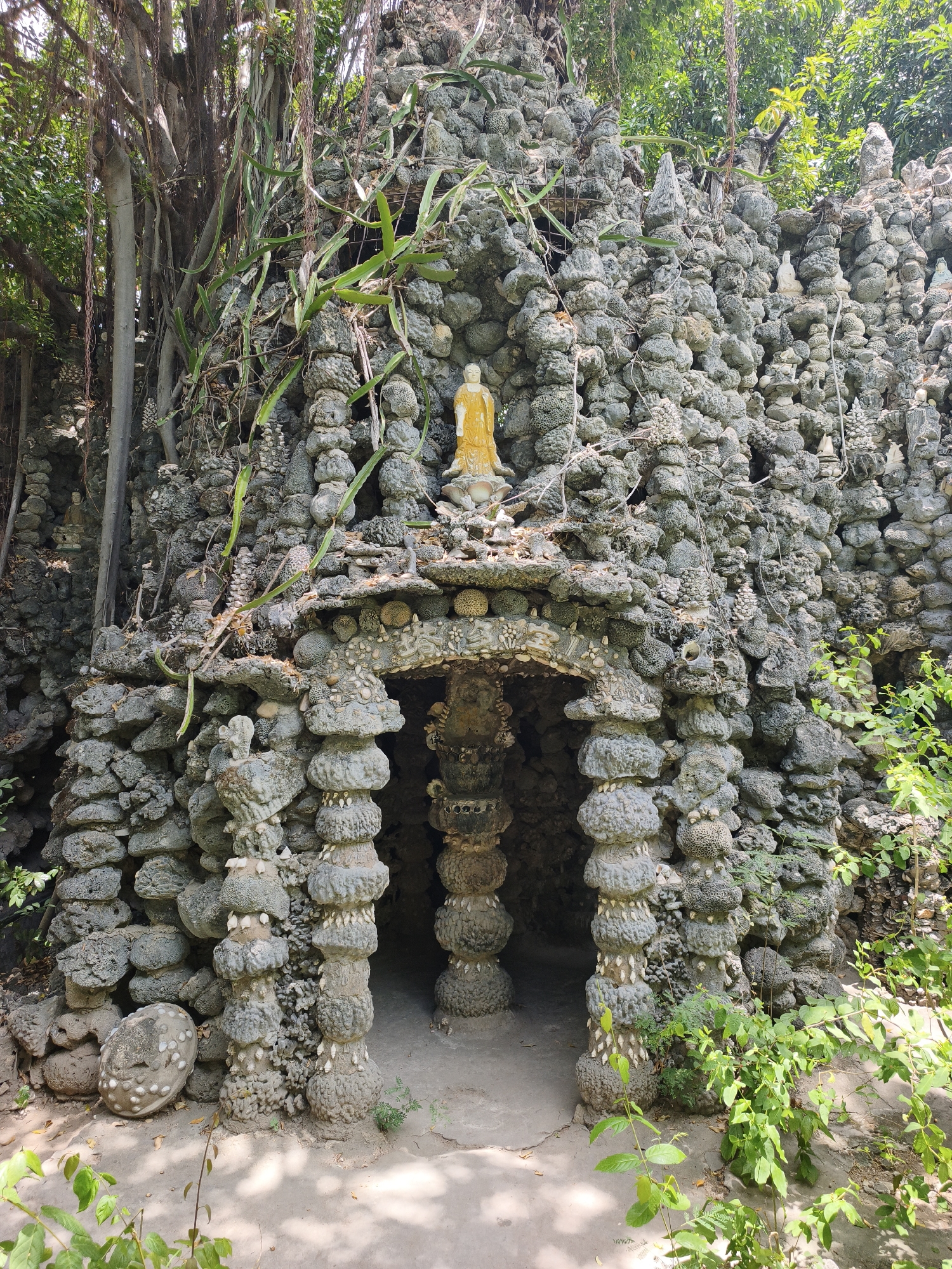 The image shows a large, elaborately constructed structure made of stones, pebbles, and shells. The structure appears to be a grotto or a shrine, with an arched entrance leading into a dark interior. A statue of a golden Buddha is positioned on top of the entrance, and smaller statues are visible throughout the facade. Lush green foliage surrounds the structure, with tree branches hanging overhead. The ground in front of the structure is paved with concrete. This unique rock garden is located at the Tu Van Pagoda in Cam Ranh, Vietnam, a Buddhist temple known for its intricate rock formations and sculptures.