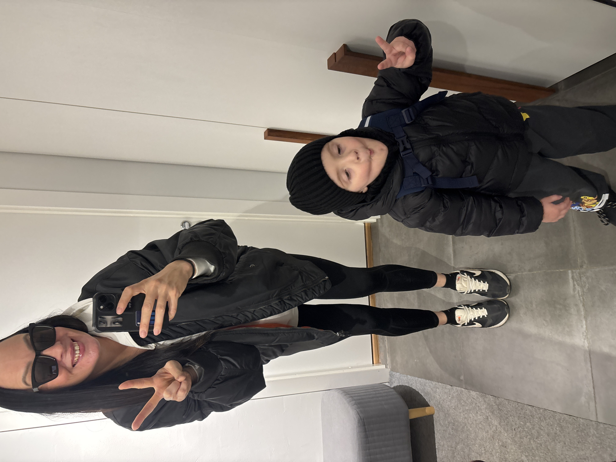 A selfie captured in a mirror shows a woman and a child posing with peace signs. The woman is wearing sunglasses, a black jacket, leggings, and sneakers. The child is wearing a black beanie, a black puffer jacket, black pants, and sneakers. The background suggests an indoor setting with white walls and gray flooring. A gray ottoman is partially visible in the lower left corner. Two wooden shelves are attached to the wall behind the child. While the location in Chaikovsky, Russia, cannot be definitively determined from this image, the objects visible do not exclude that possibility.