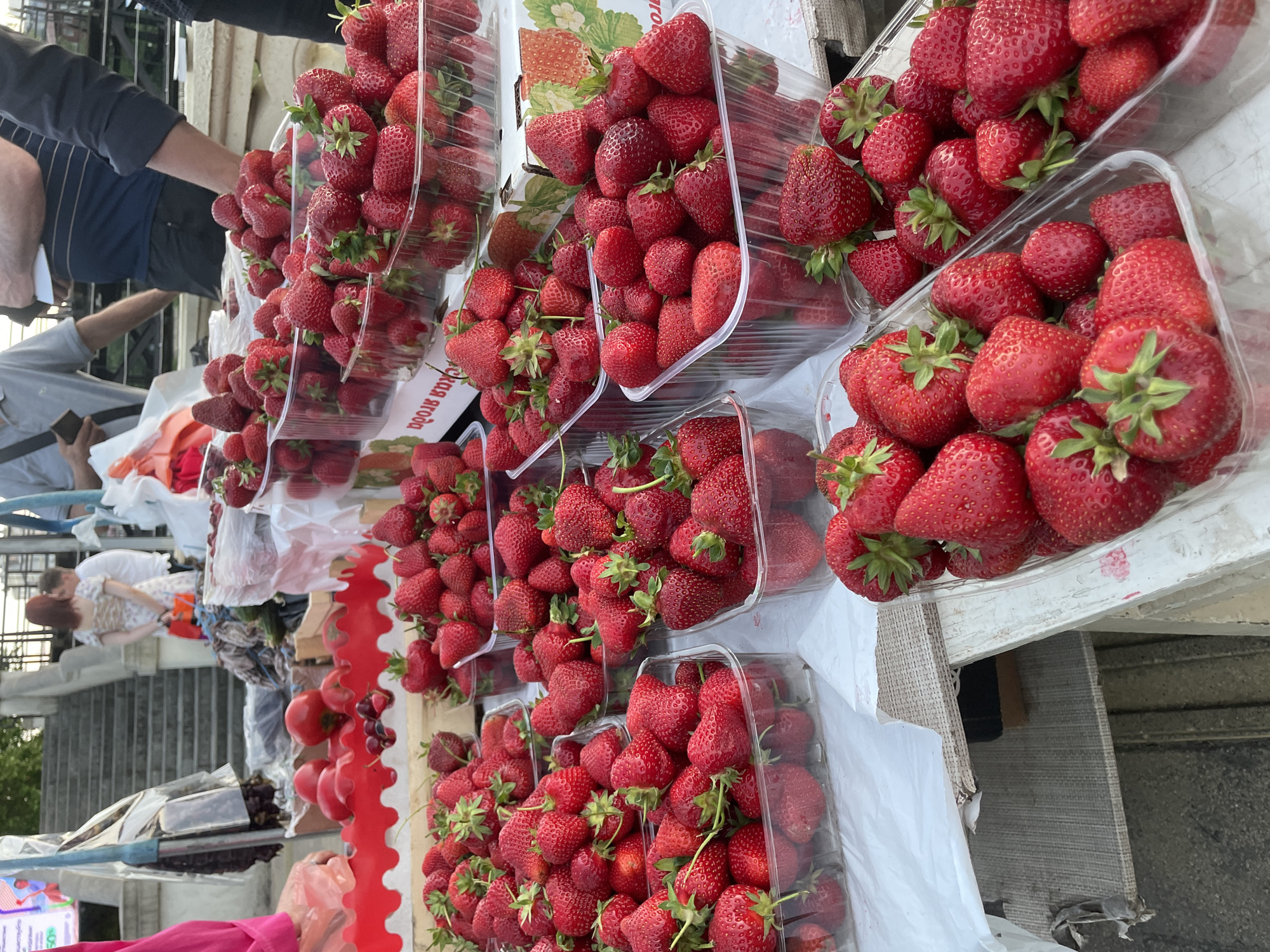 The image captures a vibrant outdoor market scene, likely in Sochi, Russia, with a strong focus on fresh produce. Numerous plastic containers are filled to the brim with bright red strawberries, piled high and glistening. These containers are arranged on what appears to be a market stall, possibly a long table or counter, with a white or light-colored surface. In the background, hints of people and merchandise suggest a bustling environment. One person's arm, clad in a blue shirt with thin white stripes, is visible reaching towards the strawberries. Another individual, wearing a white shirt, is partially seen in the mid-ground.
The scene is likely taking place during daylight hours, given the illumination and the outdoor setting. The abundance of strawberries suggests a harvest season, possibly spring or early summer. The overall impression is one of abundance and freshness, typical of a local market. The composition is dominated by the red of the strawberries and the clear plastic packaging, creating a visually striking and appetizing display. While no specific text is clearly legible within the strawberry containers, some packaging in the background might bear branding or product information, but it is too indistinct to read. The overall mood is lively and suggests a typical market day.