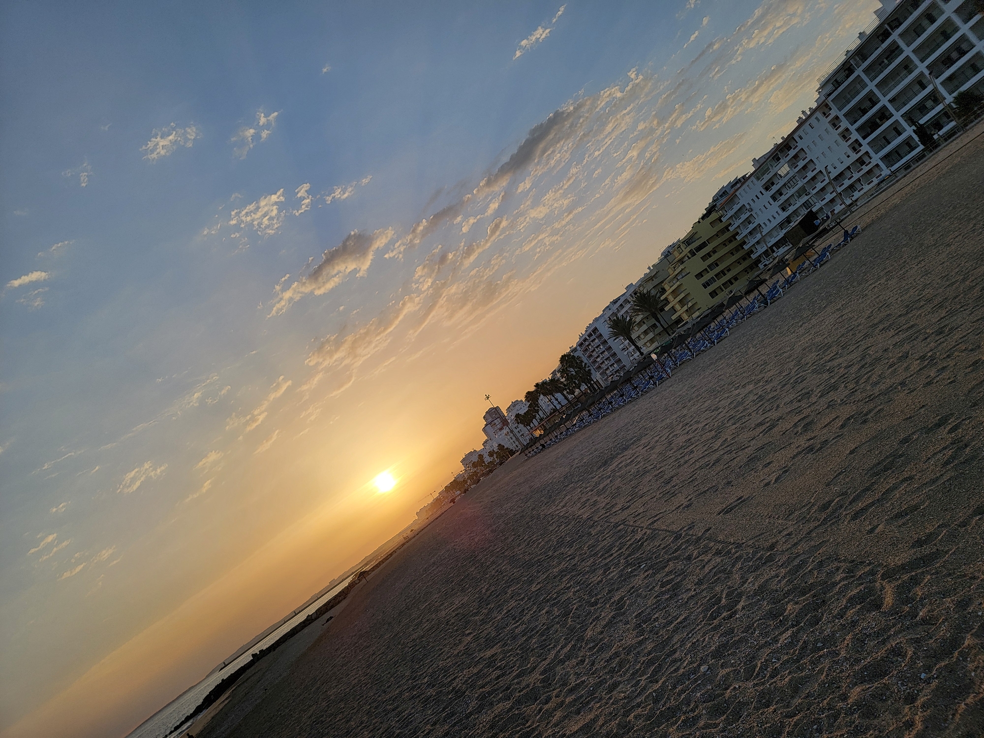 The image captures a tilted view of the Quarteira beach in Portugal at sunset. The foreground is dominated by the textured sand of the beach, leading towards the ocean on the left. The sun is a bright, warm orb low on the horizon, casting an orange glow across the sky. Wispy clouds are scattered across the blue expanse, some catching the sunset's hues. Along the edge of the beach, a line of white buildings, typical of coastal resorts, stretches into the distance, interspersed with palm trees and rows of empty lounge chairs. The scene conveys a peaceful, late-day atmosphere on a popular tourist destination.