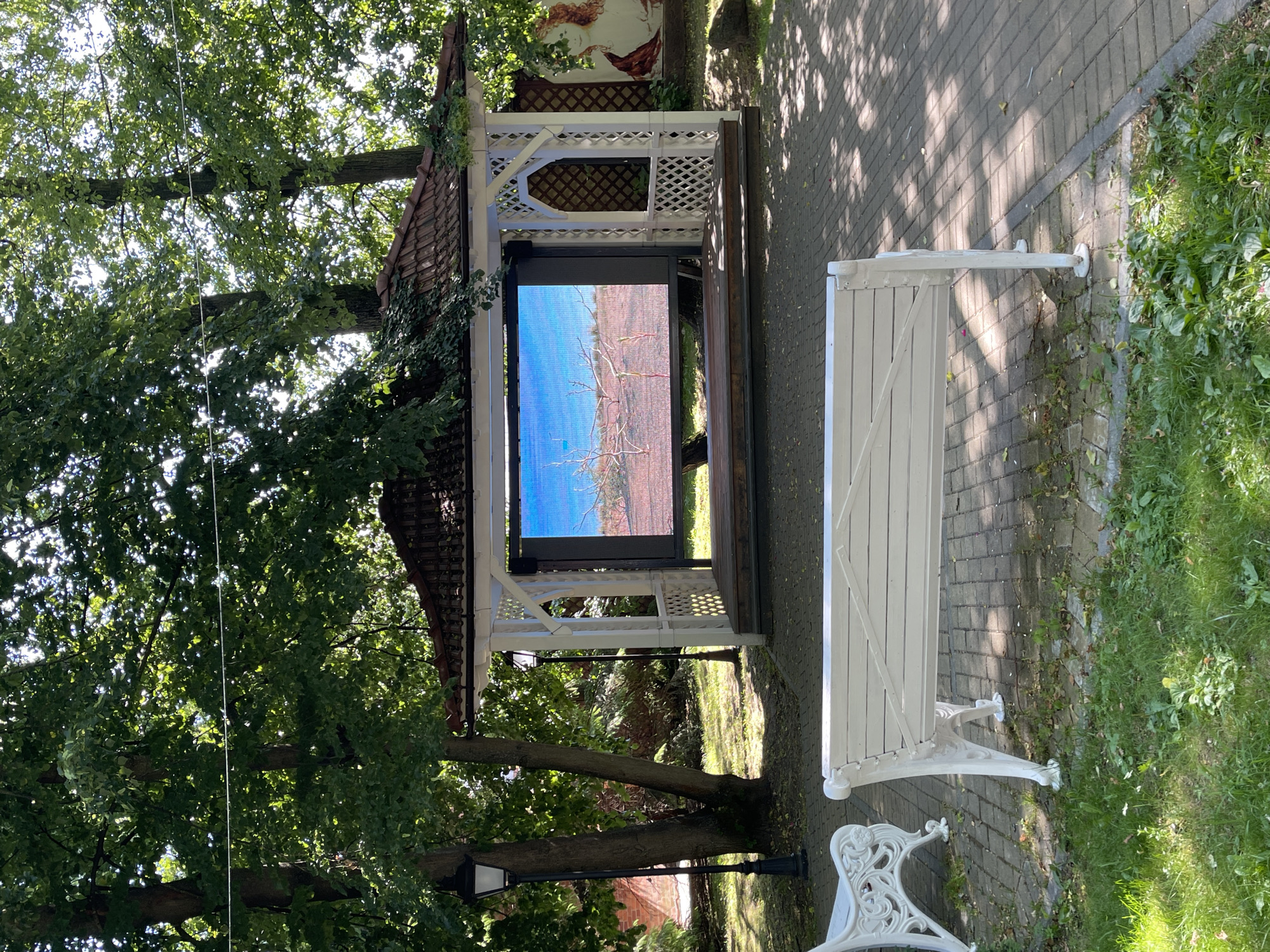 An outdoor gazebo with a large LED screen is set up in a park-like setting in Zelenogradsk, Russia. The gazebo is white with a brown tiled roof and lattice work on the sides. The LED screen displays a landscape scene with a bright blue sky and bare trees. In front of the gazebo, there are two white benches facing the screen. The ground is paved with grey brick. Tall green trees surround the area, casting dappled shadows. A lamppost stands to the left of the gazebo. In the background, a mural of two figures is visible on a wall. The scene appears to be in a public space, possibly an outdoor theater or event venue.