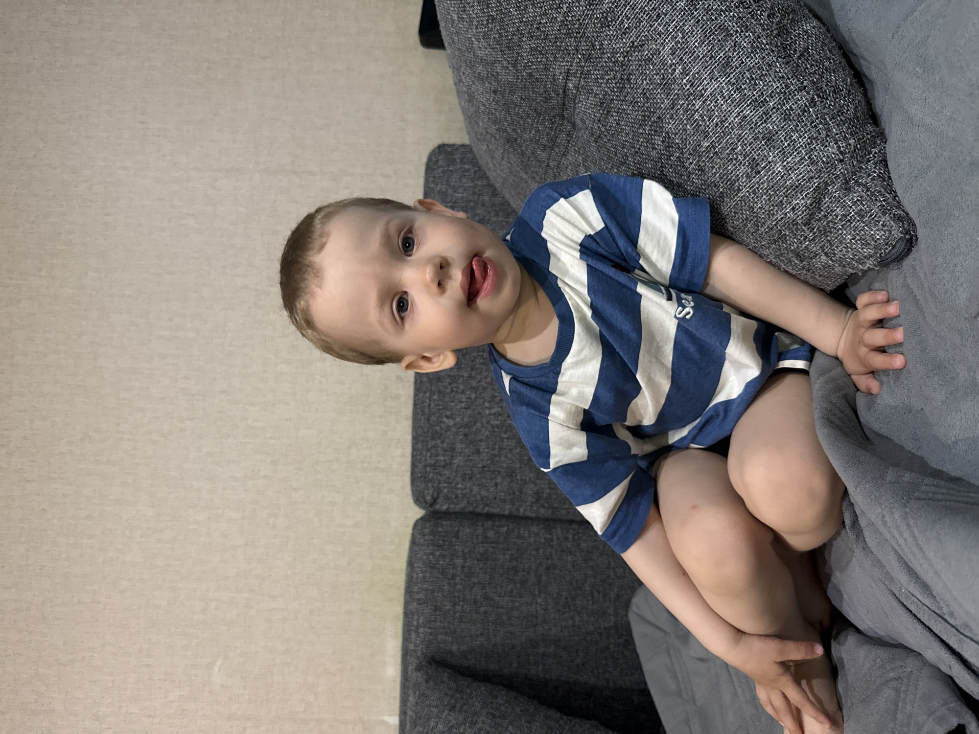 A toddler sits on a gray couch, partially covered by a gray blanket. The child wears a navy and white striped T-shirt and is looking at the camera while sticking out their tongue. The couch is positioned against a light beige wall. The location is Rusanovo, Russia.