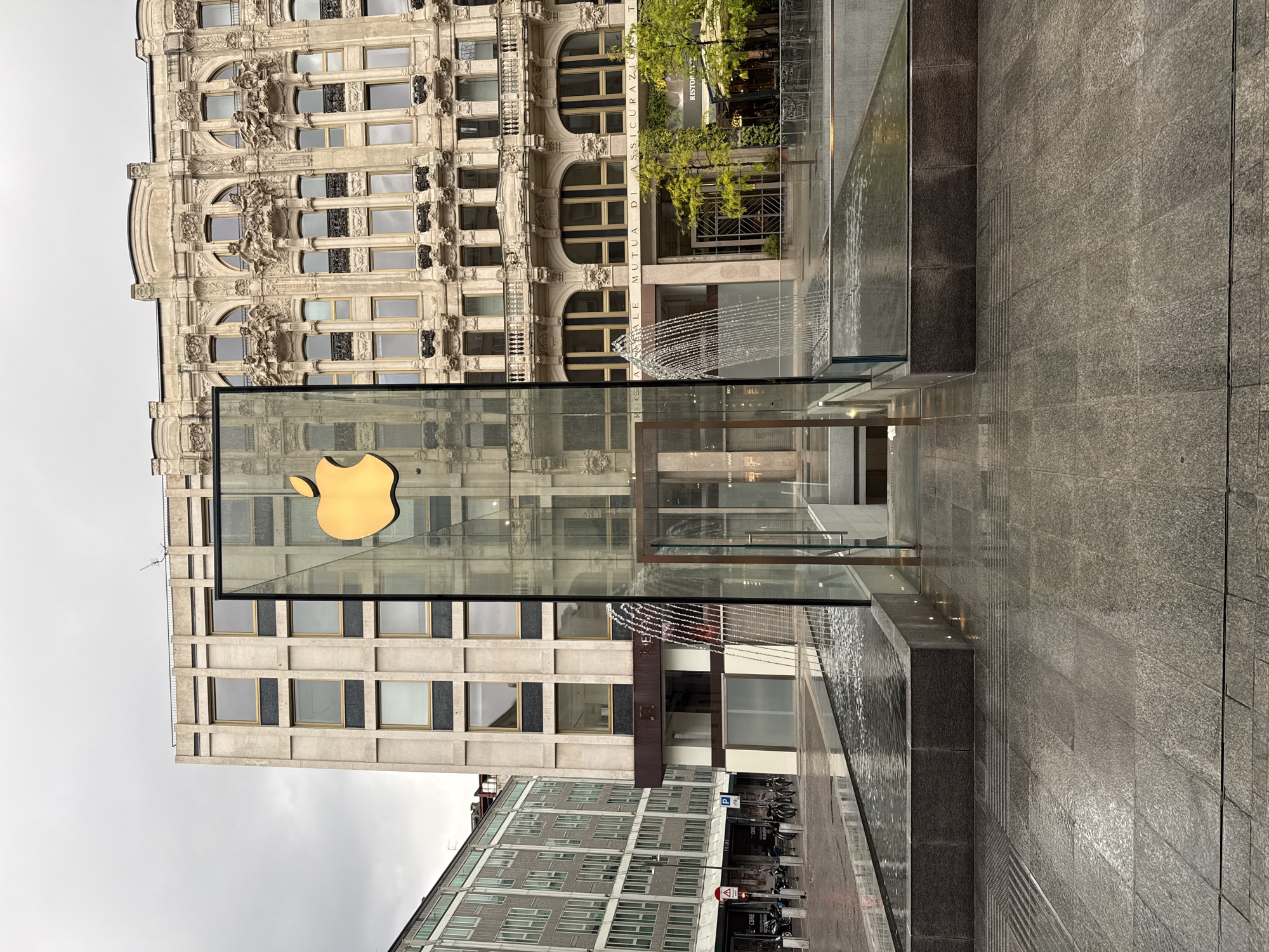 The image captures the glass cube entrance of an Apple Store, prominently featuring a large golden Apple logo, situated within an urban plaza. A modern water fountain with flowing water borders the Apple Store entrance, surrounded by dark grey paved surfaces.
In the background, two main buildings are visible. To the right of the Apple Store, an ornate historical building with intricate sculptural details, arched windows, and decorative cornices rises. To the left and slightly behind the store, a more contemporary building with multiple rectangular windows stands, and further left, another multi-story modern building with a facade of small windows is visible.
The sky appears overcast, suggesting daytime. No specific human activity is depicted, but the scene implies a commercial or public area in a city.
Visible text includes the golden Apple logo. On the ornate building, "ASSICURAZIONI" is partially legible. The words "MUTUA DI ASSICURAZIONI" are visible on another building. A sign reading "RISTORAN..." (likely "Ristorante") can be seen through some trees. Additionally, a "P" for parking and the word "CORE" are discernible on street-level signs and structures. The contrasting architectural styles are characteristic of a European city like Milan, Italy.