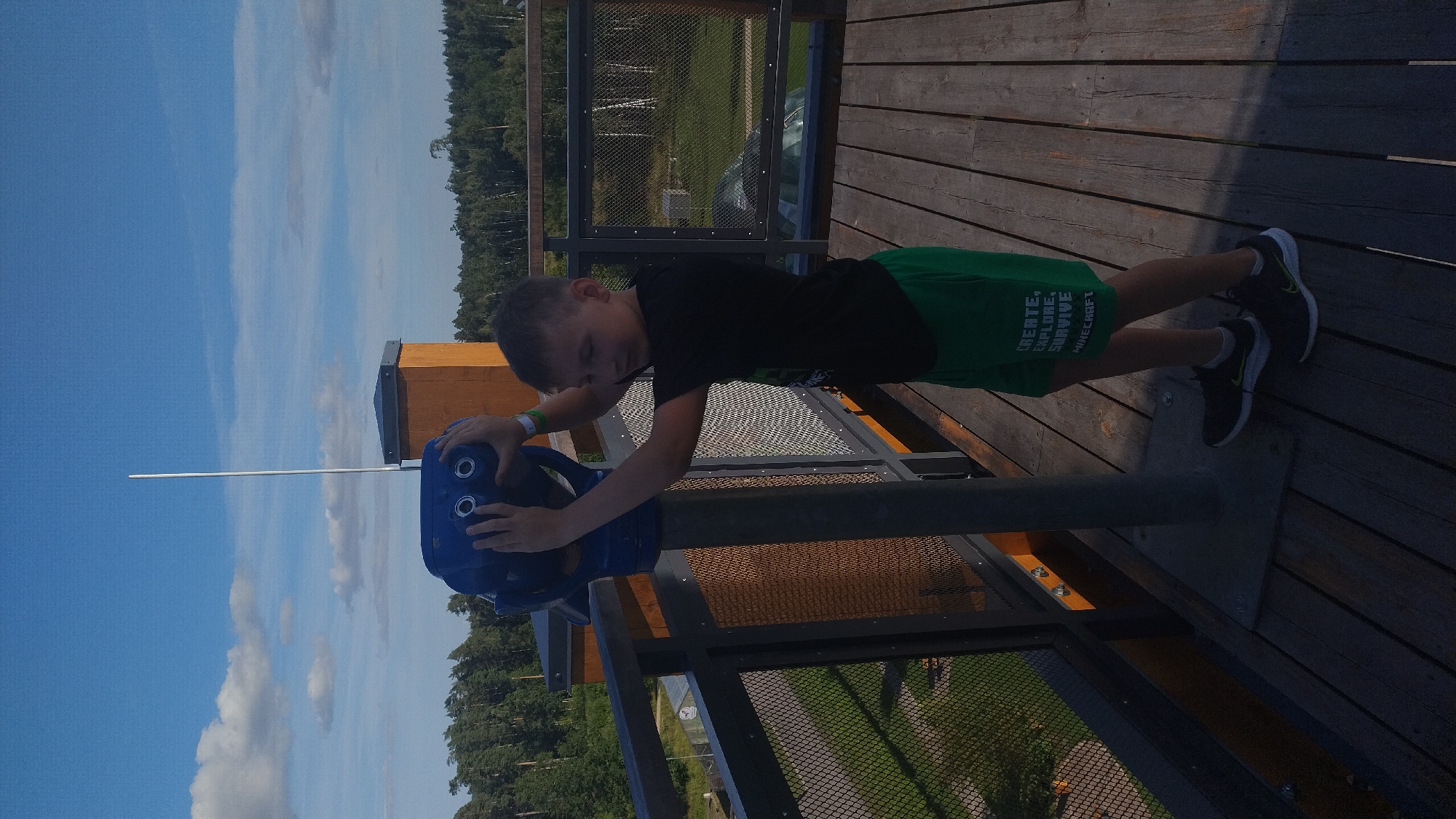 In Tērvete, Latvia, a young boy stands on a wooden platform, looking through a blue public telescope. He is wearing a black t-shirt and green shorts with the words "CREATE, EXPLORE, SURVIVE MINECRAFT" printed on them. The platform has a metal railing with a mesh infill. In the background, a dense forest stretches towards the horizon under a bright blue sky with scattered white clouds. The scene suggests a recreational area or an observation point offering panoramic views of the surrounding landscape.
