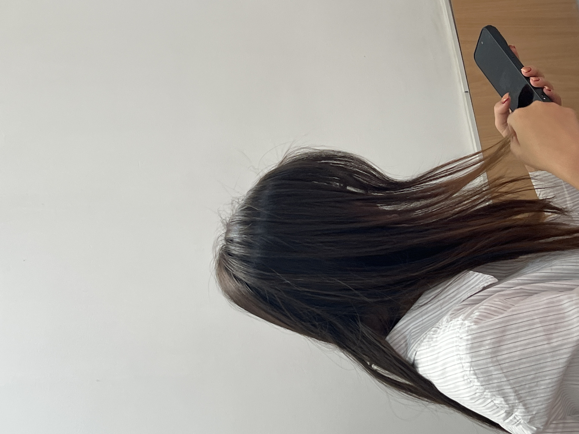 A person, with long, dark brown hair, is shown from the chest up, in profile, facing right and looking down at a black smartphone they are holding in their hands. They are wearing a white collared shirt with thin, vertical grey pinstripes. The background is a plain white wall, with a wooden trim line visible horizontally across the lower portion of the image. The scene appears to be indoors, possibly a room or an office. There is no visible indication of the specific location within the city of Kadamjay, Kyrgyzstan.