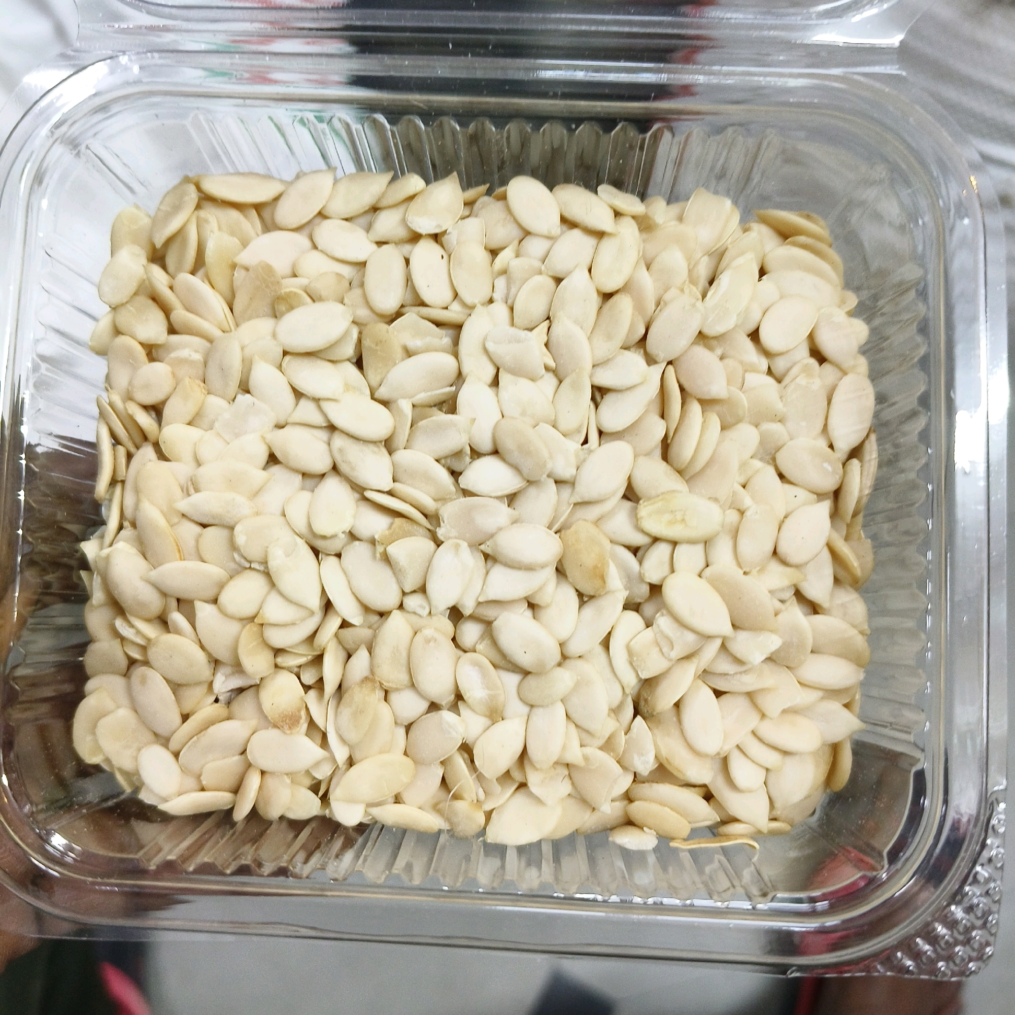 A close-up, overhead shot shows a clear plastic container filled with a large quantity of light-colored, oval-shaped seeds. The seeds appear to be watermelon seeds, some with a small point at one end and a rounded tip at the other. They are packed tightly together, creating a textured surface within the container. The plastic container has a ribbed texture along its base and clear, smooth sides. The background is blurred, but appears to be a light, possibly reflective surface. There are no visible indications of location in Karimnagar, India, or any people, actions, or specific environmental details beyond the object itself.