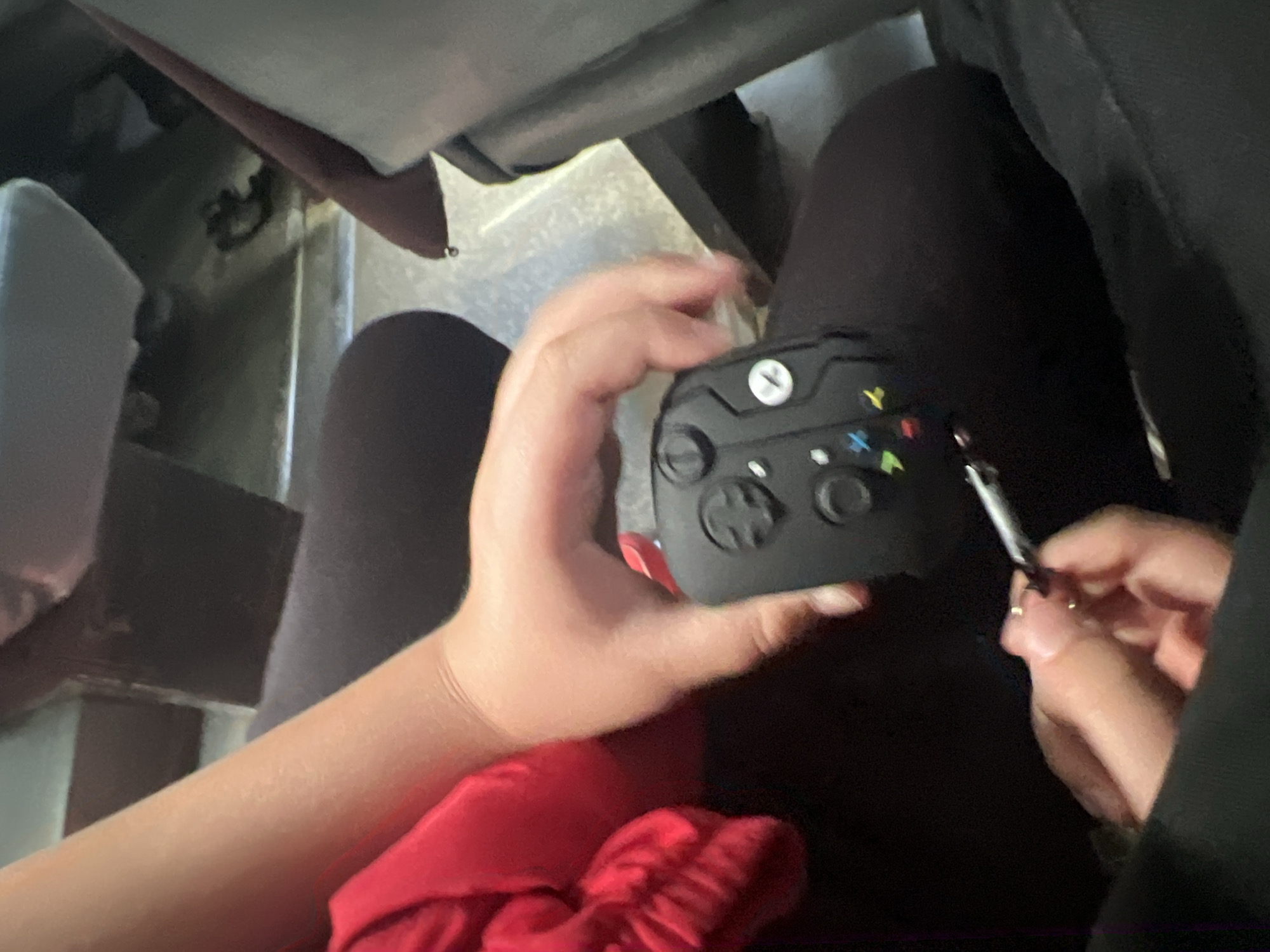 The image depicts a close-up view of hands interacting with a black video game controller, possibly an Xbox controller, which is resting on dark fabric, likely clothing. One hand is holding the controller, and another hand is manipulating a small, silver object, which appears to be a joint or cigarette. The background is blurred but suggests an indoor setting with some furniture visible. There are no discernible landmarks or clues to place the image in Ulyanovsk, Russia, or any specific city. The overall lighting is dim.