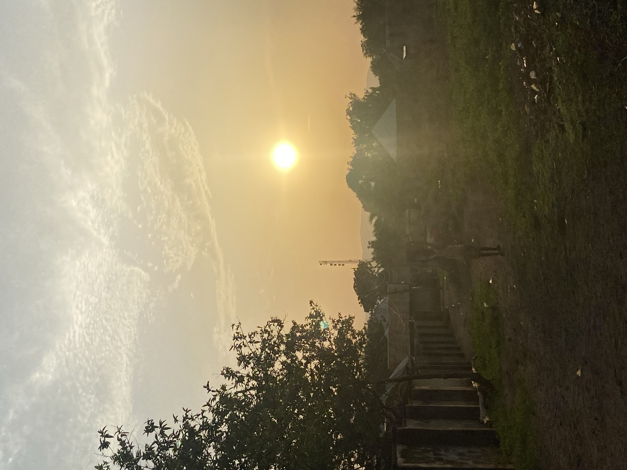 The image shows a partially obscured view of Mararaba, Nigeria, at sunset or sunrise. The sun is visible in a hazy sky, partially obscured by clouds and smog. In the foreground, the silhouette of a tree is visible. To the right, concrete steps lead upwards beside a mossy or vegetation-covered wall. A construction crane is also visible in the distance. Two people are walking away from the camera on a path. The air quality appears poor.