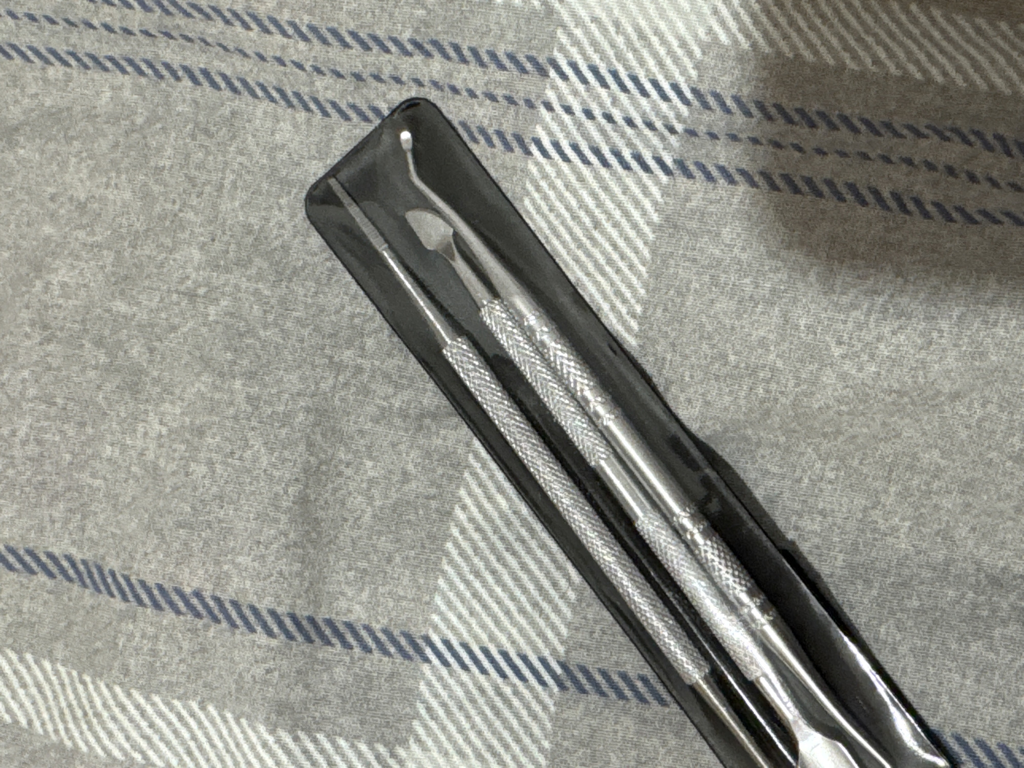The image shows a set of three dental or carving tools in a clear plastic case, resting on a grey fabric surface with a blue and white plaid pattern. The tools appear to be made of metal and have textured handles for grip. The case is open, revealing the tools within. The lighting suggests an indoor setting. No people or actions are visible. The fabric surface is consistent with bedding or upholstery.