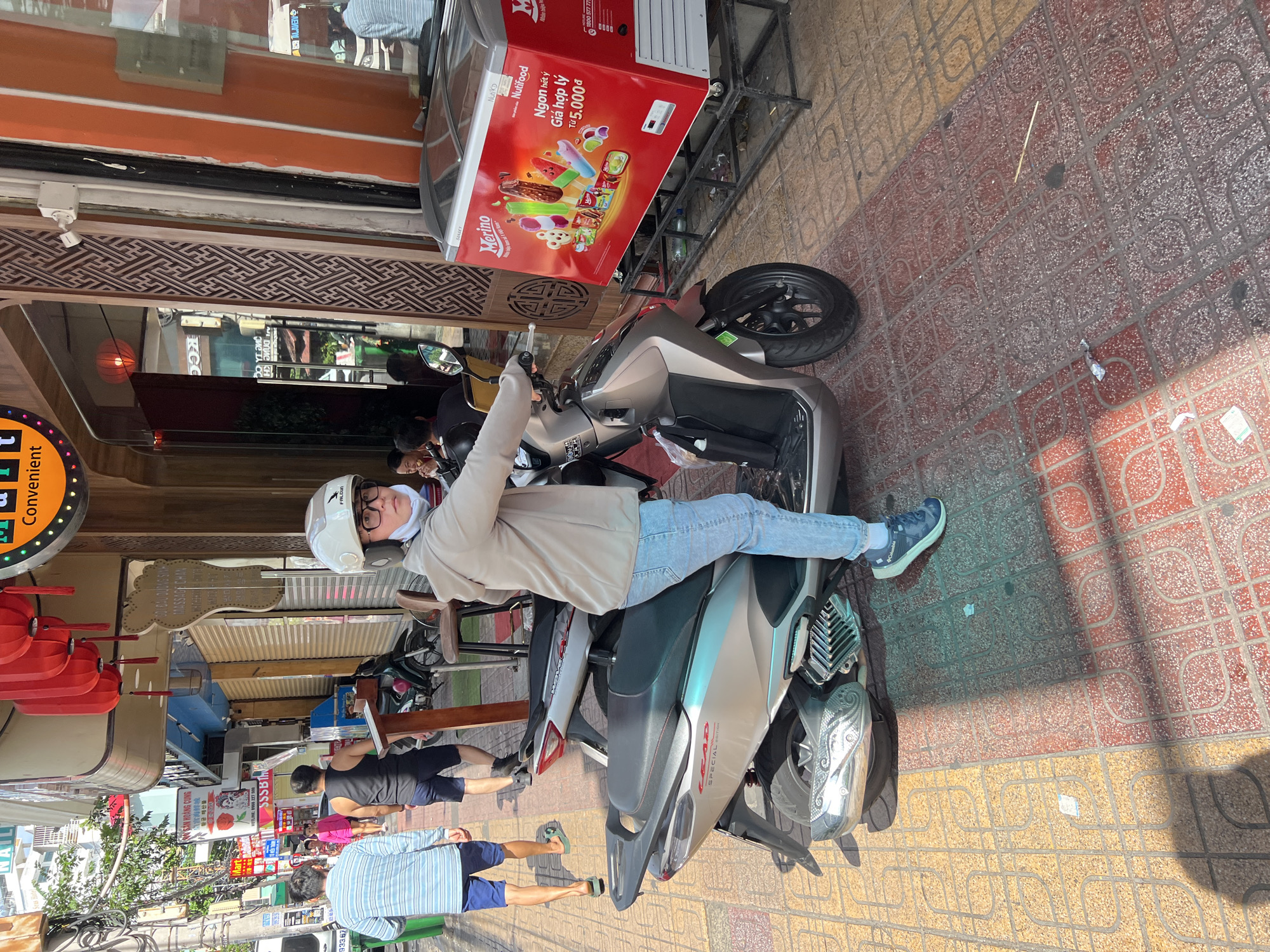 The image depicts a street scene in Nha Trang, Vietnam. In the foreground, a person wearing a white helmet and a light-colored hooded jacket is seated on a silver scooter. The person is wearing blue jeans and dark sneakers. The scooter is parked on a sidewalk with a patterned tile floor. To the right of the scooter is a red freezer filled with ice cream. In the background, other people can be seen walking on the sidewalk and a variety of shops line the street. Red lanterns hang from an awning above one of the shops. The overall scene suggests a typical day in a Vietnamese city.