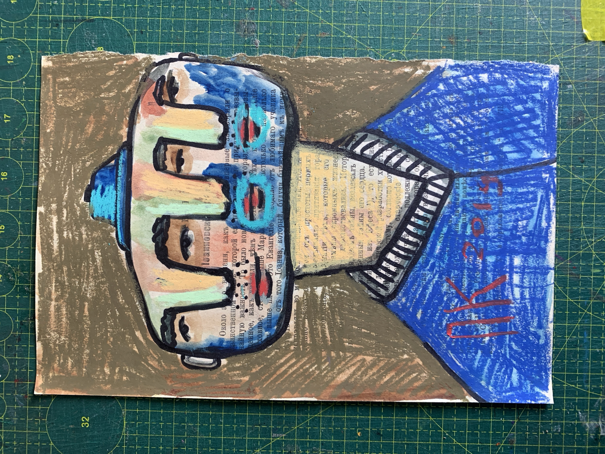 The image is a photograph of a piece of art, possibly a mixed-media collage or painting on paper. The artwork features a stylized depiction of a multi-faced head wearing a blue hat, rendered in various colors including blue, yellow, green, and flesh tones. The background of the artwork appears to be newspaper clippings or text. Red markings, possibly a signature or date, are visible on the lower portion of the artwork, appearing to read "ПК 2019". The artwork itself is placed on a green cutting mat with a grid pattern. There are no elements in the image that provide information about the city of Sergeevo, Russia.