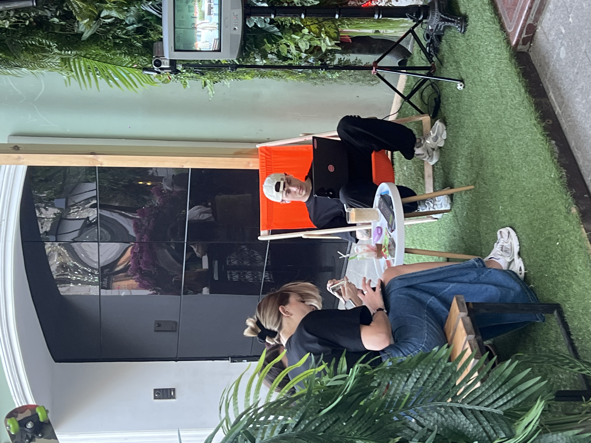 This image depicts a semi-outdoor seating area in the city of Moscow, Russia. The scene features two individuals engaged with electronic devices, alongside various objects and a video monitoring setup. On the left, a light-skinned female with light brown hair tied in a high messy bun with a black scrunchie sits on a wooden slatted bench with a black metal frame. She wears a black short-sleeved t-shirt and a long, faded blue denim skirt, paired with white New Balance sneakers. Her head is bowed, and she is actively using a smartphone held in both hands, with another device resting on her lap. Behind her, a white arched doorway frames a large, dark, reflective facade composed of multiple panels. Reflections within the facade include distorted light sources, what appears to be purple floral arrangements, and interior elements. Two dark grey intercom units are mounted on the white wall adjacent to the dark facade. A camouflage-patterned skateboard with green wheels is propped against the upper left side of the archway. In the center, between the two individuals, stands a small, round white table with three wooden legs. On the table are a tall glass with a light brown beverage and a straw, a shorter glass with a clear liquid and a straw, a black smartphone, a white rectangular case (possibly earbuds), a small clear pouch with colorful items, and a black device. On the right, a light-skinned male with a white baseball cap worn backward sits in an orange fabric deck chair with a wooden frame. He wears a black short-sleeved t-shirt and black pants, with light grey sneakers. He is looking down and using a black laptop resting on his lap, which has a small pink circular sticker on its lid. Earbuds are visible in his ears. To his right, a dense wall of various green plants, predominantly ferns, extends vertically. Positioned in front of the plant wall is a video recording setup consisting of a silver camcorder mounted on a black tripod. Below the camcorder, a silver Samsung CRT-style monitor displays a live feed of two figures in white garments standing outdoors in what appears to be a courtyard. Cables are visible on the ground around the tripod base. The ground surface of the seating area is covered with artificial green turf, which abuts a grey concrete or stone paved area in the immediate foreground. A light green wall is