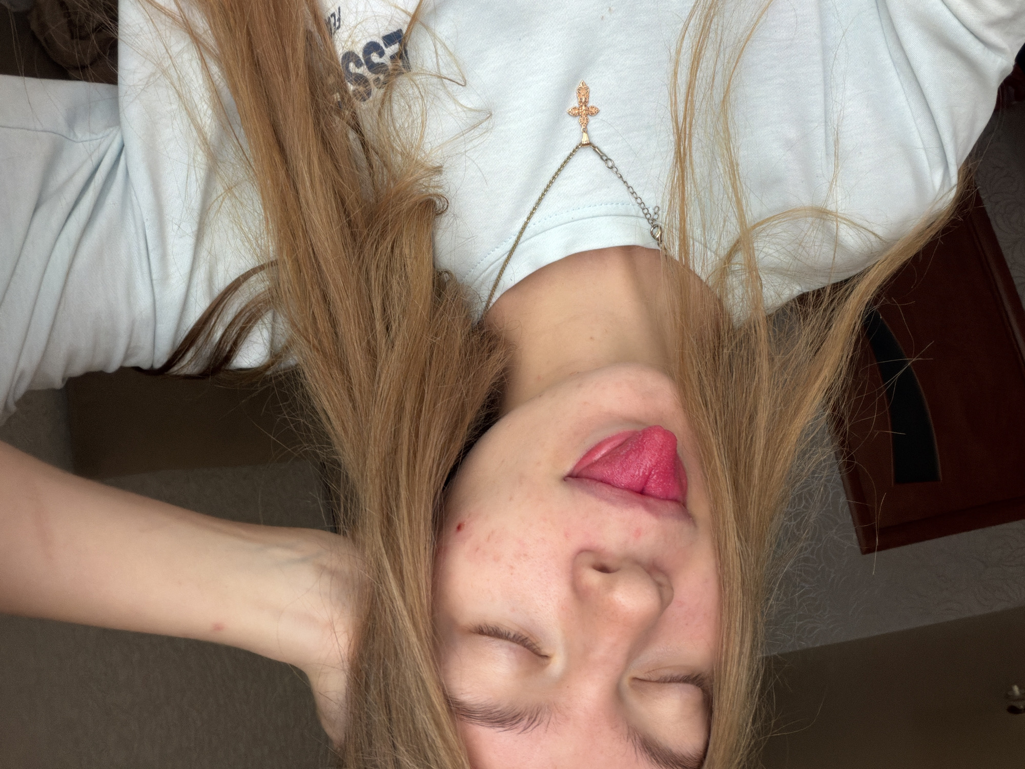 In a medium shot, a young woman with long, light brown hair pulled back on one side of her head is seen with her eyes closed and her tongue sticking out. She is wearing a light blue t-shirt with a pendant necklace featuring a cross. A red mark is visible on her cheek. The background appears to be an indoor setting with a wooden door to the left and a textured wall. The lighting is dim, suggesting it may be indoors during evening hours. The image does not contain any elements that would suggest a specific location within the city of Chaikovsky, Russia.