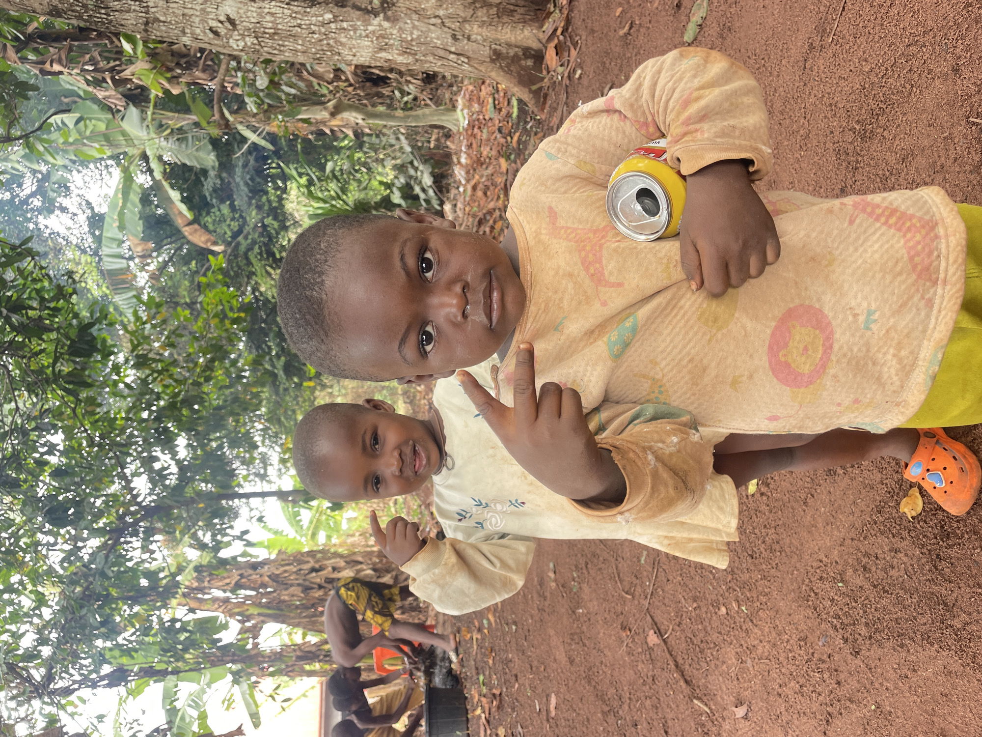 Two young children stand on a red dirt ground in Ogbe, Nigeria. The child in the foreground holds an opened yellow can and gestures with their fingers. The second child is slightly behind and to the left, also gesturing with their fingers. Both children wear long-sleeved, patterned shirts. The child in the foreground wears orange footwear. In the background, other individuals are present near a water source. Lush greenery surrounds the scene. A large tree trunk is visible at the top left of the frame.