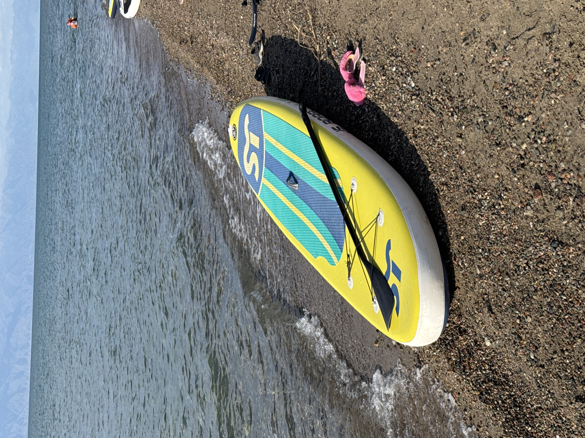A yellow stand-up paddleboard with a blue and green striped design is resting on a pebble beach next to a body of water. A black paddle is placed on top of the paddleboard. In the background, mountains are visible across the water under a cloudy sky. A person is partially visible in the water in the distance, wearing an orange and white life vest. A pair of pink fuzzy slippers are lying on the beach near the paddleboard. Two black shoes are also on the beach behind the paddleboard, along with a dark object that appears to be a paddleboard handle or accessory. The scene is likely in Basbeshik, Kyrgyzstan, given the geographical features.