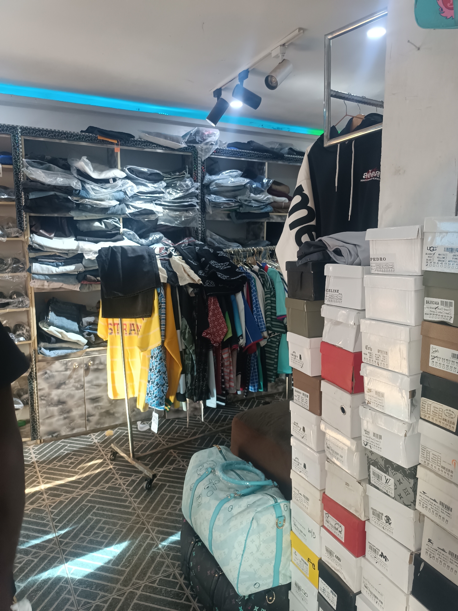 The image depicts the interior of a clothing and footwear store in Bukuru, Nigeria. Shelves are laden with folded clothes and garments on hangers. A rack displays various articles of clothing. Stacks of shoe boxes bearing brand names like "UGG", "Balenciaga", "Celine", "Pedro", "Gucci", and "Prada" are visible. A blue patterned travel bag sits near the shoe boxes. Overhead are track lighting fixtures. A person wearing a black t-shirt is partly visible in the foreground.