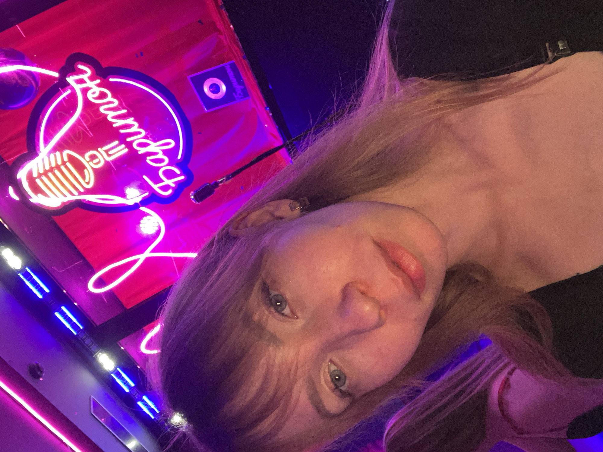 A woman with fair skin and shoulder-length light brown hair is taking a selfie in what appears to be a bar or entertainment venue in Perm, Russia. She is positioned slightly off-center, with her head tilted. In the background, there's a neon sign that reads "The Bagman" with a microphone design. The signage includes a logo bearing Cyrillic text. The lighting in the venue is dominated by purple and pink hues, likely from LED strips or neon lights. The venue appears to be a nightclub or bar called “The Bagman” in Perm, Russia.