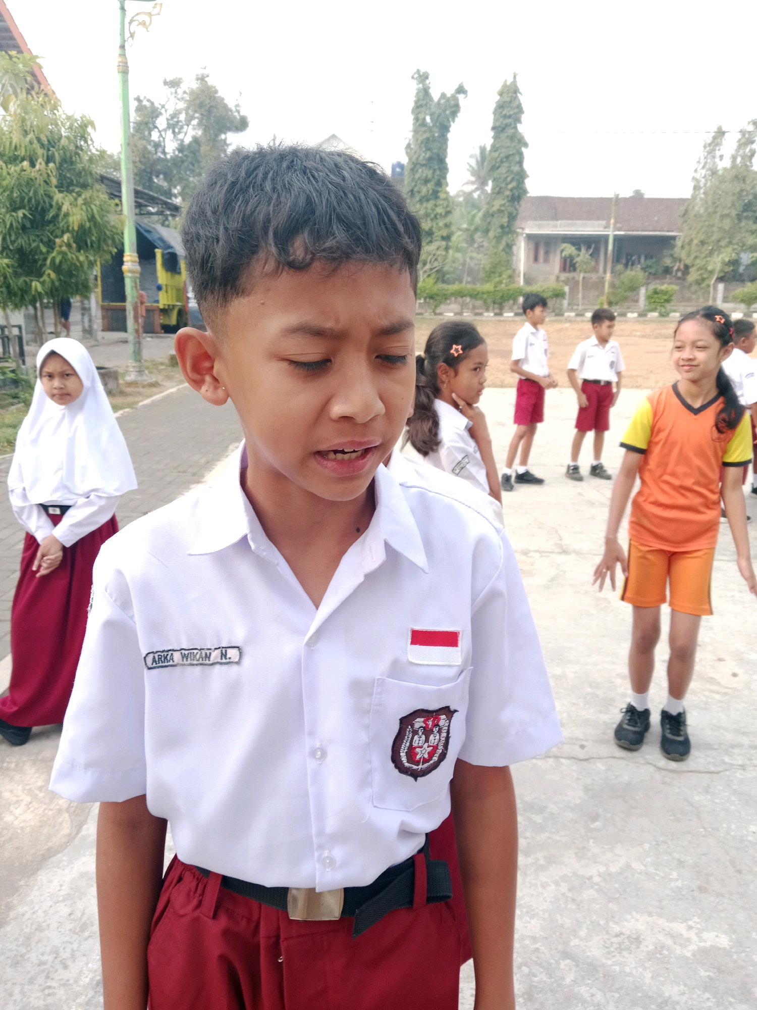 The image shows a close-up of a young boy in the foreground wearing a white school uniform shirt with red trim on the pocket and a nametag that reads "ARKA WIKAN N.". He is also wearing red pants and a black belt. Behind him, several other children in school uniforms are visible. To the left, a girl in a white hijab and red skirt stands looking towards the camera. In the background, there are trees, buildings, and a light pole. The scene appears to be an outdoor schoolyard. The location is identified as Salatiga, Indonesia.