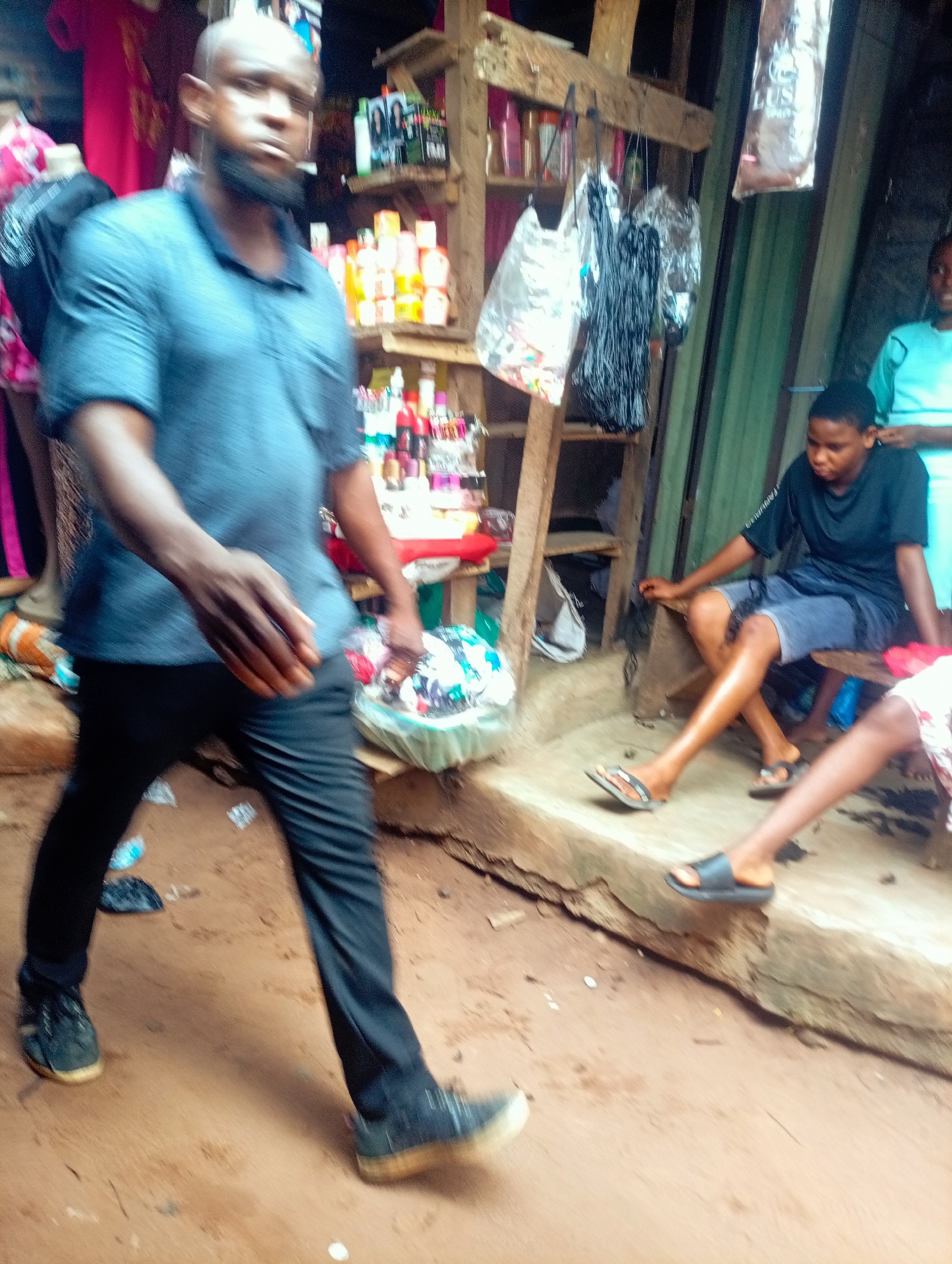 In Nkpor, Nigeria, a man with a shaved head and a beard, wearing a dark blue collared shirt and black pants, walks towards the viewer on a dirt path. He is in motion, with his right arm slightly extended and his left arm bent at the elbow. Behind him, to his right, a young woman with short hair, wearing a black t-shirt and blue shorts, sits on a wooden bench, looking down and to her left. To the right of the young woman, a person wearing a red and white patterned top is partially visible, with their legs extended. In the background, a market stall is set up with wooden shelves displaying various items, including bottles, bags, and packages. The environment appears to be an outdoor market with a rough, uneven ground.
