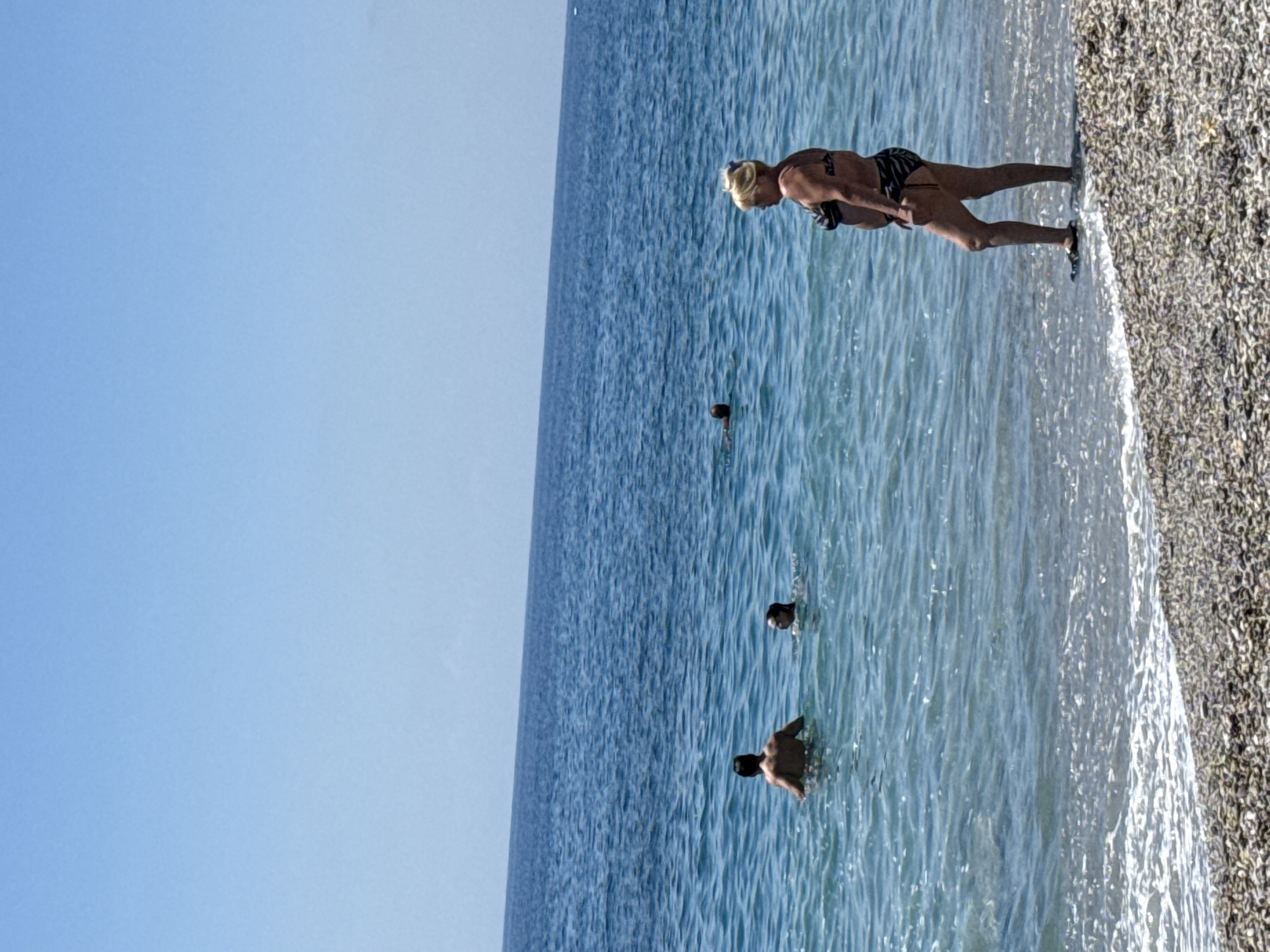A wide-angle shot depicts a beach scene. The sky is light blue and clear. The ocean is blue with gentle waves. Along the shoreline, there is a pebble beach. A woman in a bikini is standing at the edge of the water near the pebble beach. Three people are swimming in the ocean at varying distances from the shore. The city and specific location are not discernible from the image.