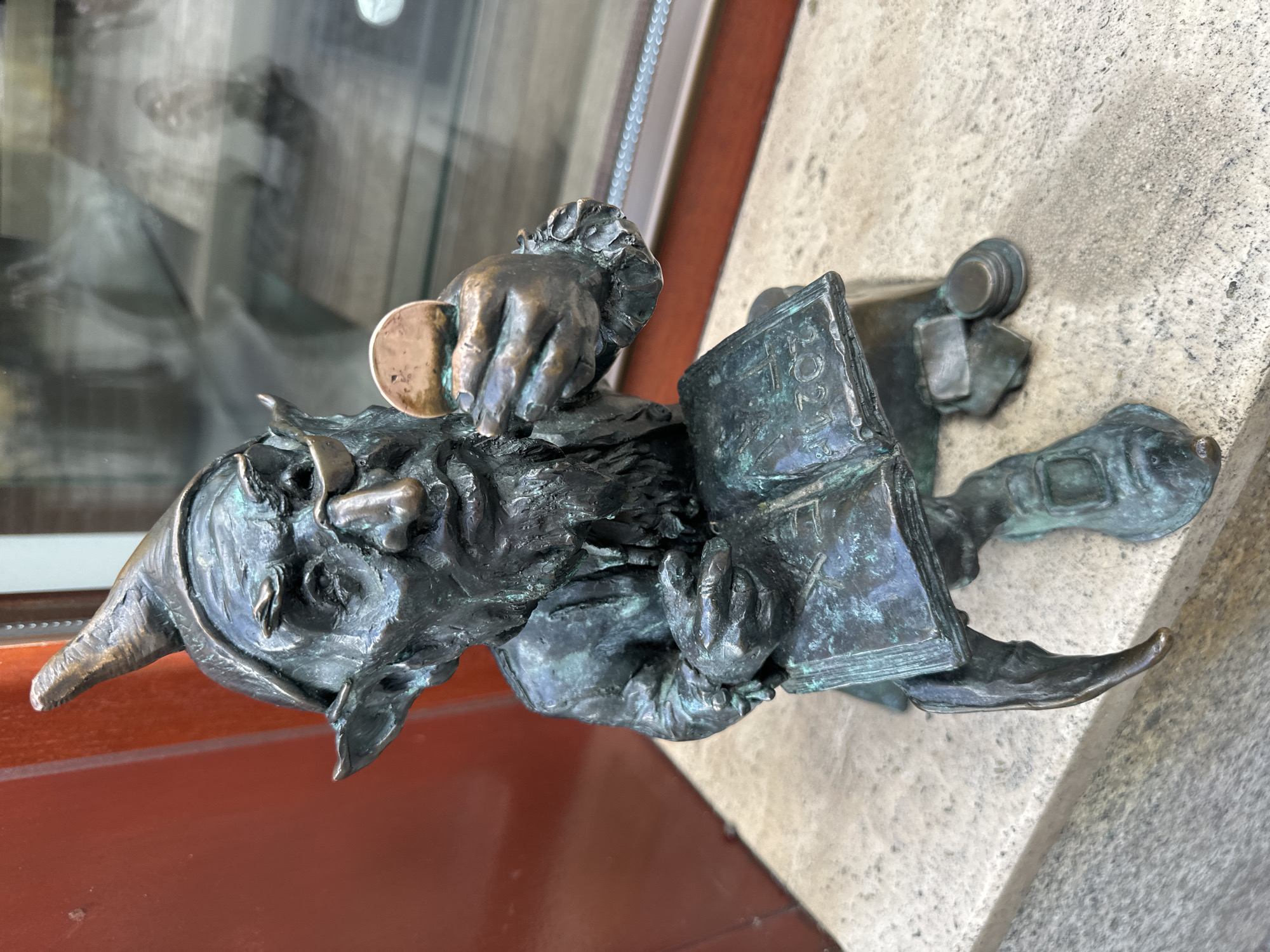 A bronze statue of a gnome in Wrocław, Poland, affixed to a building’s exterior. The gnome wears a pointed hat and is holding a book with the year "2021" inscribed on its cover. It is also holding a coin. The gnome appears to be in mid-stride, with its right foot partially raised. The statue is positioned on a stone ledge, with a window visible in the background.