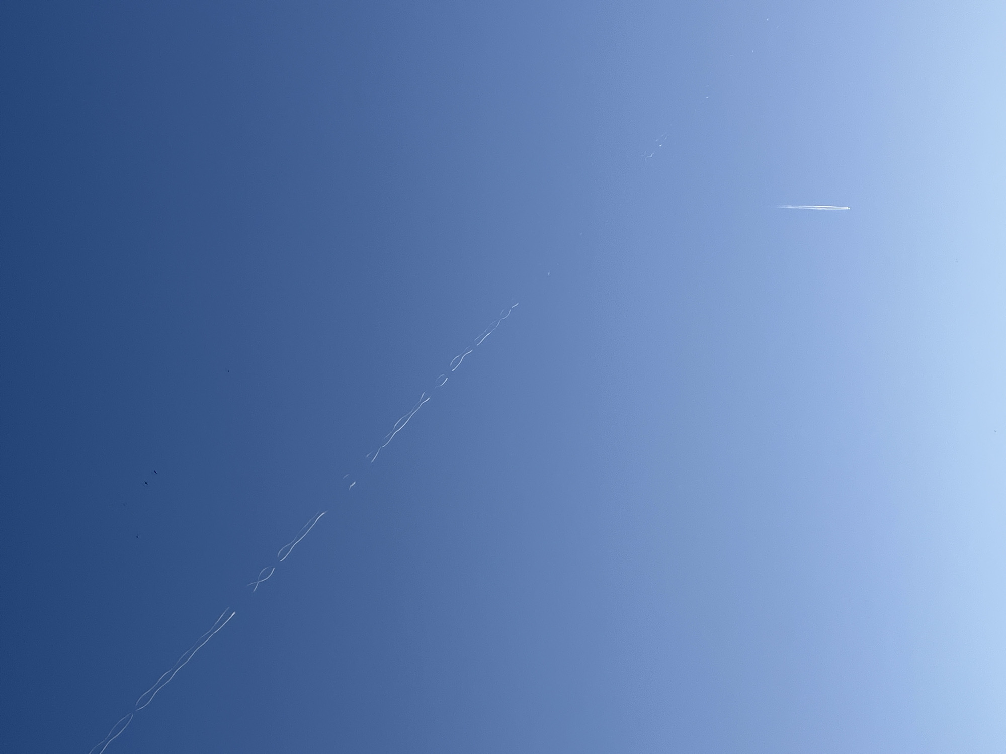 A clear, blue sky fills the frame. Several contrails of airplanes are visible. One contrail is relatively straight and short, located in the upper right quadrant. Another contrail is longer and more sinuous, appearing as a dotted line, descending from the upper center towards the lower left corner of the frame. A small cluster of dark objects, possibly birds, is visible in the lower left quadrant.
