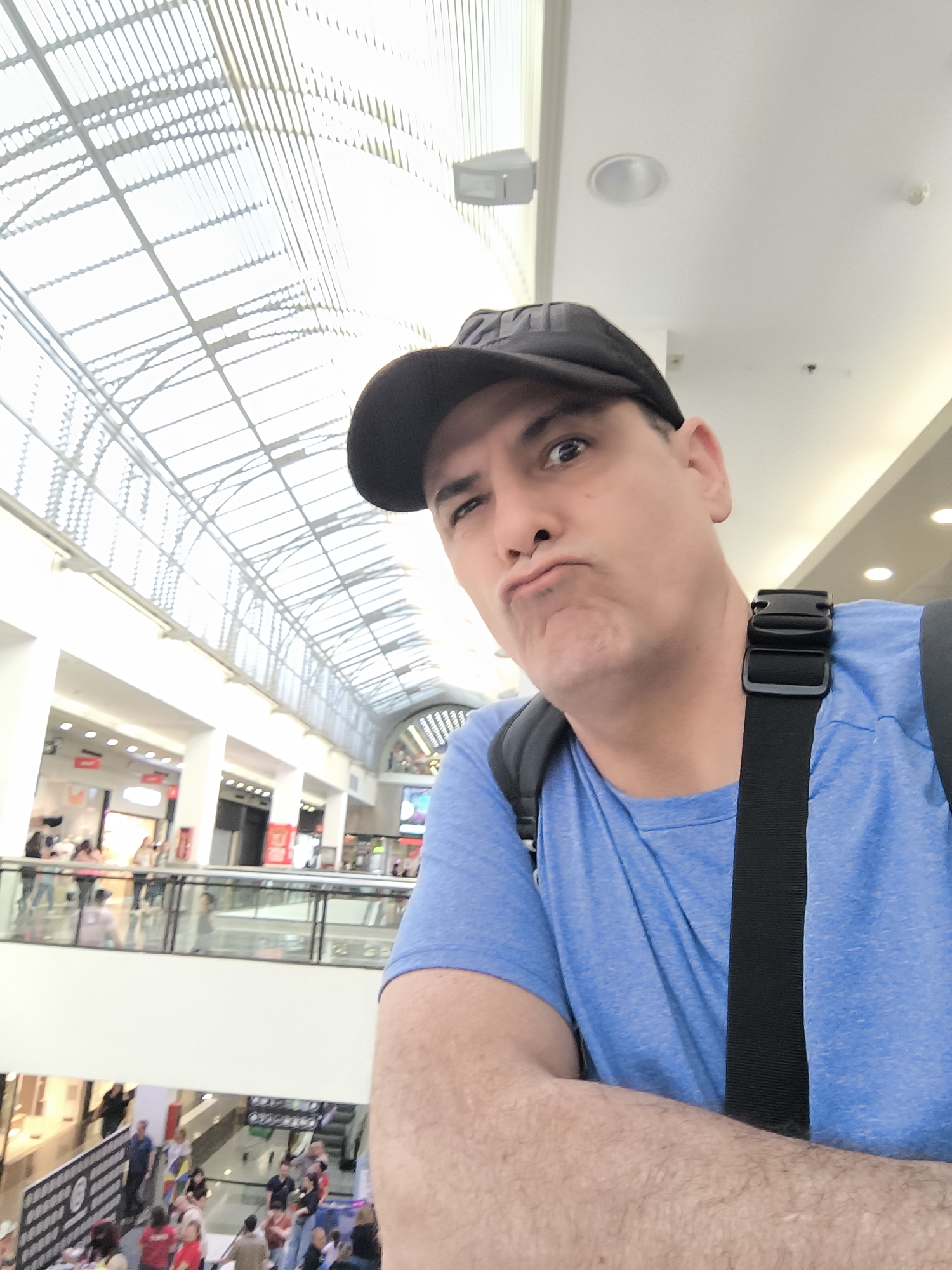 This image is a selfie taken by a man making a playful, pouting expression, looking directly at the camera. He is wearing a black baseball cap, a bright blue t-shirt, and a black backpack with a visible strap across his right shoulder. His right arm is bent and resting in front of him.
The setting is a large, brightly lit indoor shopping mall in Barberà del Vallès, Spain. The mall features a striking arched glass ceiling with a white grid-like metal framework, allowing ample natural light to illuminate the space, suggesting it is daytime. Below the ceiling, multiple levels of the mall are visible, characterized by white walls, pillars, and glass railings. Numerous people can be seen in the background on different floors, walking and congregating, indicating a busy commercial environment. Storefronts with various displays line the upper levels, and recessed lighting fixtures are integrated into the architecture. The text "HM" is faintly visible on the front of the man's cap, but no other specific text on distant signs is clearly legible.