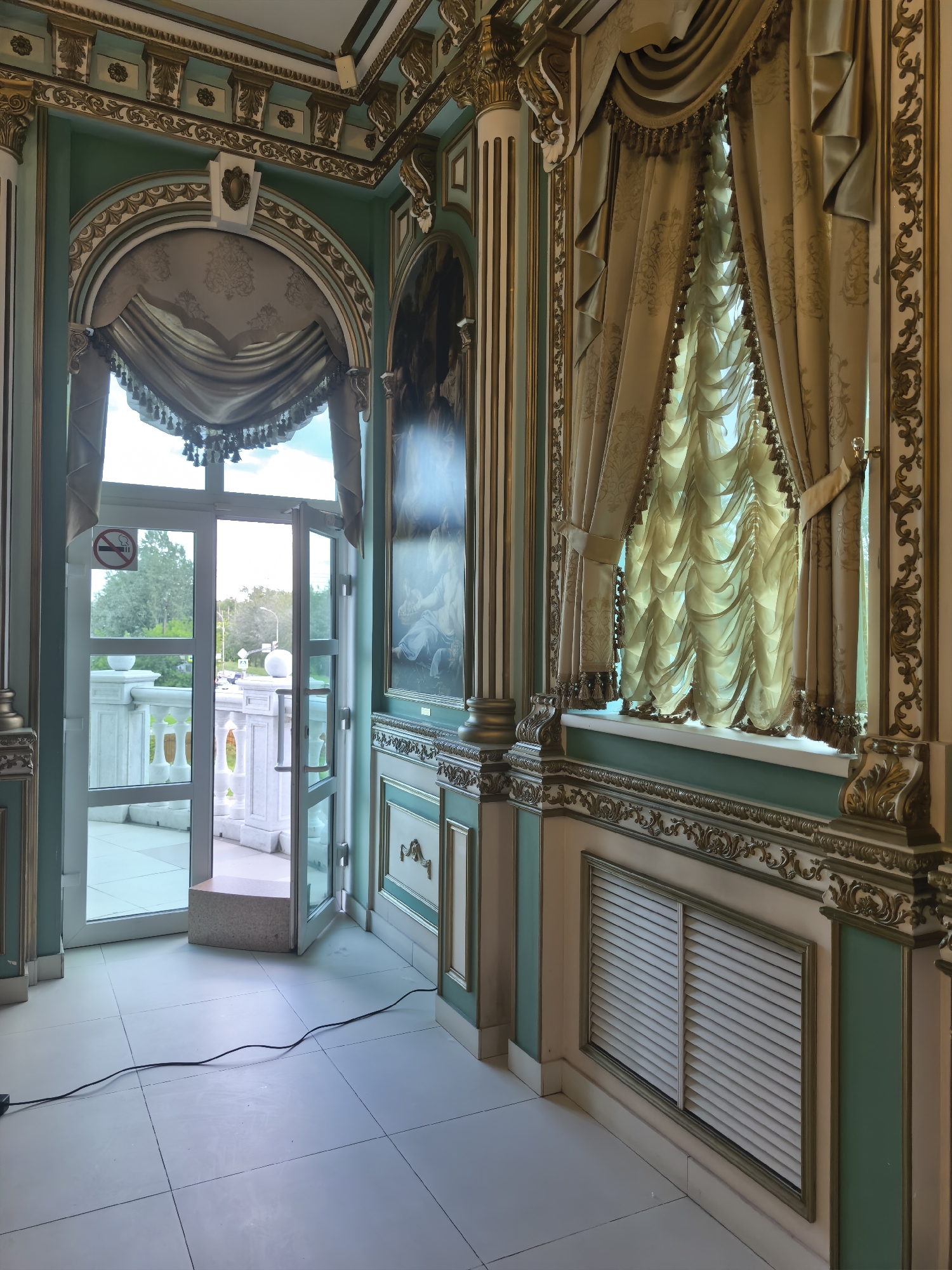 An interior view shows a richly decorated room with ornate gold trim, light green walls, and large windows with elaborate gold curtains. A set of open glass doors leads to an outdoor patio with a balustrade. A black electrical cord runs across the tiled floor. The room appears to be a lobby or common area within a grand building. There are no people visible in the image.