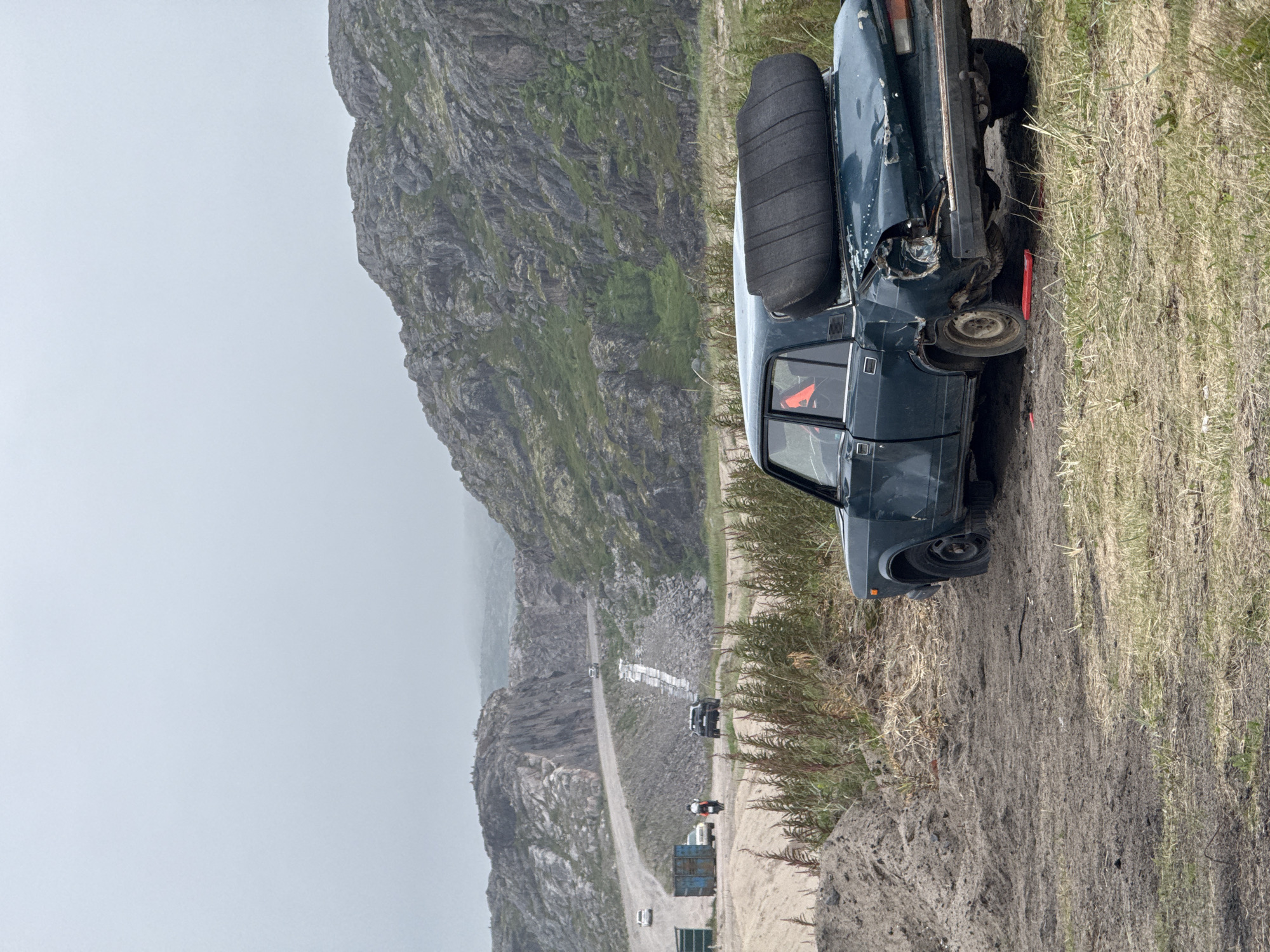 The image depicts a desolate, rocky landscape near Teriberka, Russia, characterized by a dirt road winding through hills and sparse vegetation. In the foreground, a dark green, heavily damaged sedan is parked off-road. The car's rear is crushed, with a flat tire visible on the left rear wheel. A dark, cylindrical object, possibly a rolled-up mattress or blanket, is precariously balanced on the trunk. A red triangular warning sign is visible inside the car's rear window. In the middle ground, several vehicles, including a motorcycle, a black SUV, and two white cars, are present on the dirt road. Further in the distance, a large blue shipping container sits by the side of the road. The sky is overcast, contributing to the somber atmosphere of the scene, which appears to be a quarry or a remote industrial area.