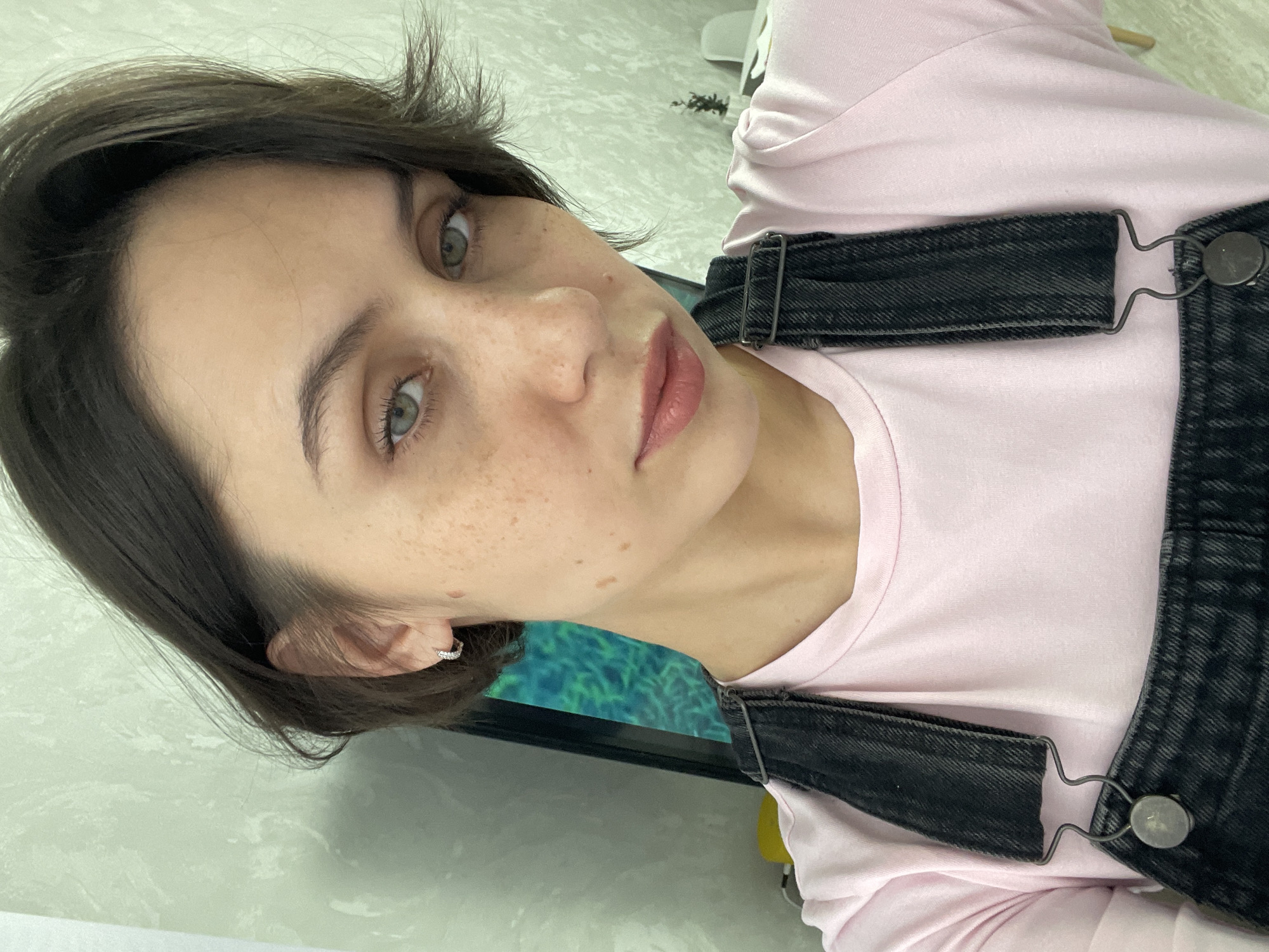 The image is a portrait-style close-up of a young woman, rotated 90 degrees clockwise. She has short, dark brown hair that is styled and appears slightly tousled. Her striking eyes are a light greenish-blue, and her face is marked with numerous freckles across her nose and cheeks. She wears a soft, natural-looking pink lipstick and has prominent eyebrows and long eyelashes. A small, shiny hoop earring, possibly adorned with tiny stones, is visible in her left ear.
She is dressed in a light pink long-sleeved t-shirt, layered under dark gray or black denim overalls. The overalls feature metal buckles and fasteners on the straps, one of which is visible over her right shoulder. Her expression is neutral and direct as she looks into the camera.
The background suggests an indoor setting, possibly a home or office. A light-colored, subtly textured wall or surface occupies much of the upper left and background. To her right, behind her shoulder, a dark rectangular object, likely a monitor or screen, displays a vibrant abstract pattern of green and blue hues, resembling foliage or an underwater environment. The lighting is soft and even, typical of indoor conditions. There is no visible text in the image. The general location context is Izhevsk, Russia.