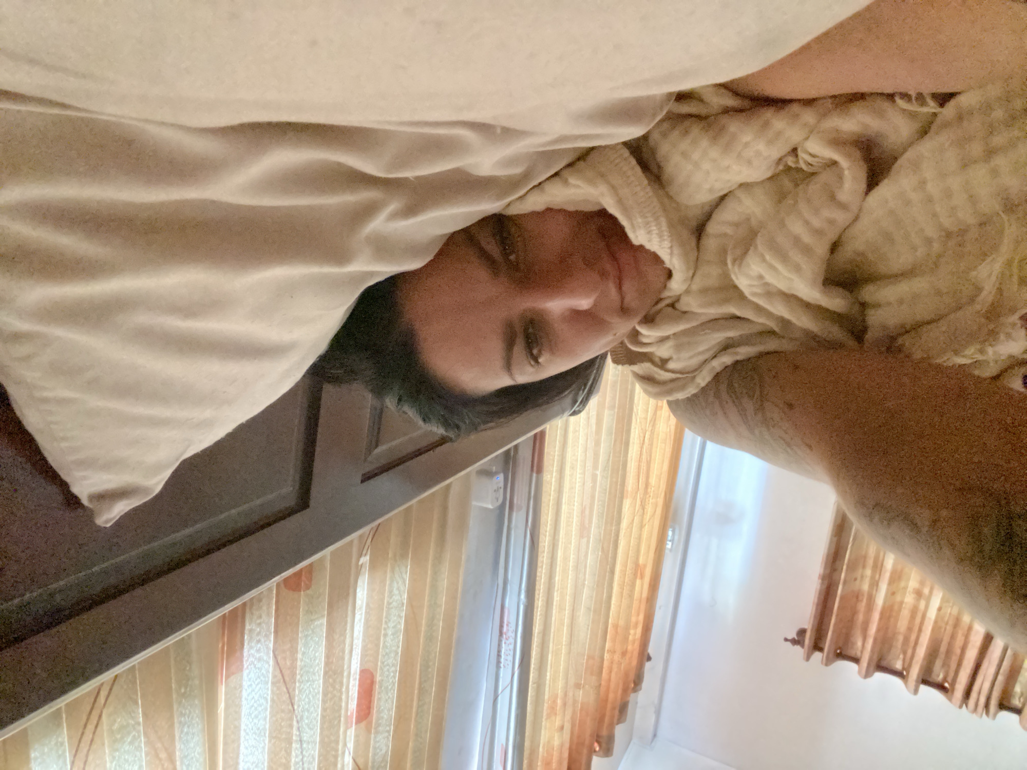 A person lies in bed, facing the camera, under a light-colored blanket. The person has dark hair and is looking towards the camera with a slight smile. The background shows a wooden headboard and sheer curtains. The overall scene suggests a relaxed or intimate setting within a bedroom. The image does not provide any specific clues about the city of Mũi Né, Vietnam.
