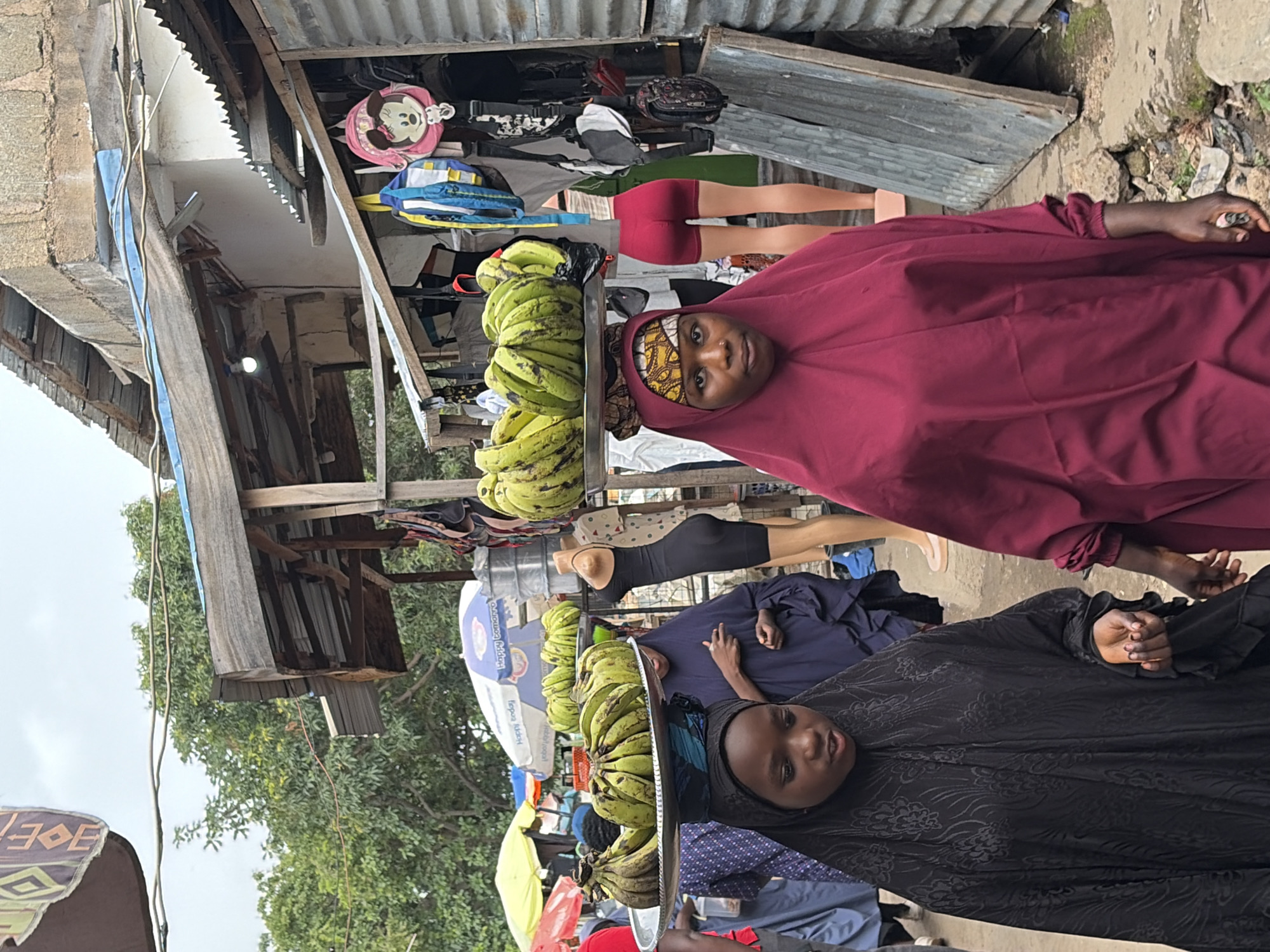 In Jos, Nigeria, a busy outdoor market scene unfolds. Two young women are the central focus, both wearing hijabs and long, modest clothing. The woman on the left, dressed in a black garment with a subtle pattern, carries a large metal tray laden with bunches of green bananas balanced on her head. Her eyes are directed downwards, and her right hand is slightly raised, fingers open. To her right, another woman, adorned in a deep maroon hijab and matching clothing, walks alongside. She looks directly at the camera, her left hand holding a cigarette. Behind them, other market stalls are visible, with corrugated metal roofs and wooden structures. A headless mannequin wearing a black, form-fitting dress and red shorts stands to the right of the women. Various bags, including a pink one with Minnie Mouse on it, hang from a stall in the background. Umbrellas, one yellow and another blue with the text "Happy Tomorrow," provide shade in the cluttered marketplace. The ground is rough, with scattered rocks and debris.